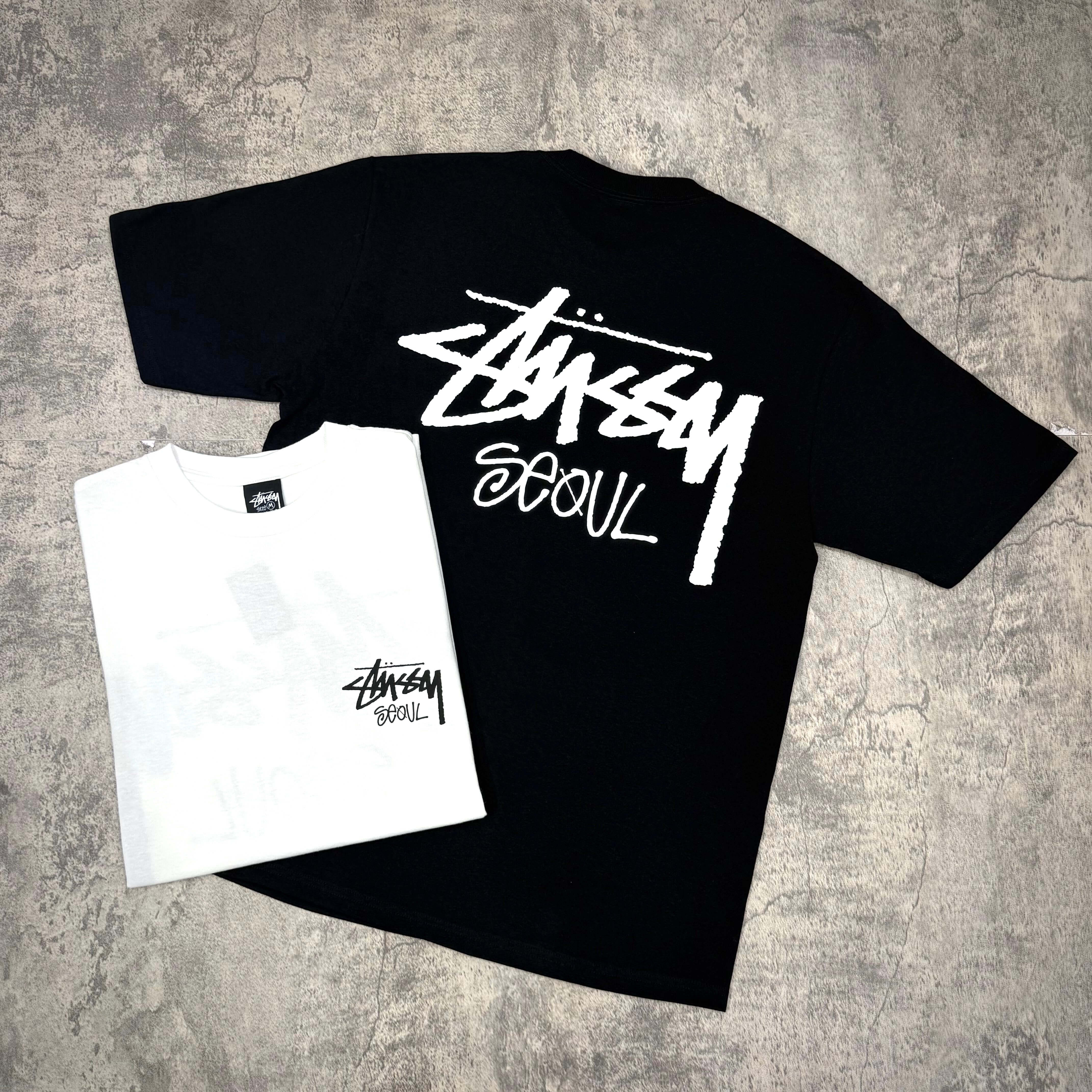 Stussy Seoul Exclusive Logo Tee in black and white laid flat on a textured surface.