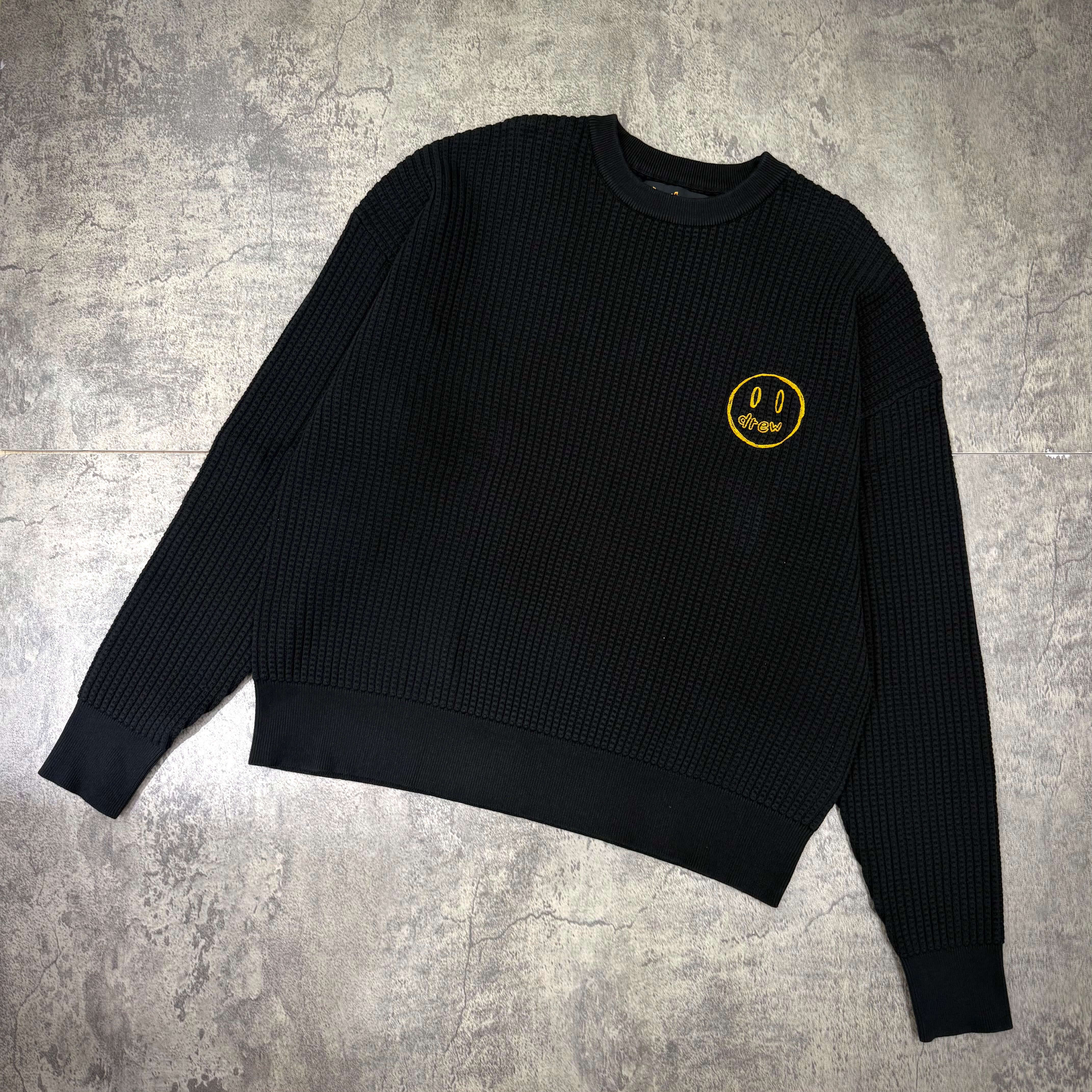Drew House Mascot Waffle Sweater - Black with yellow smiley logo on the chest, displayed flat on a textured surface.