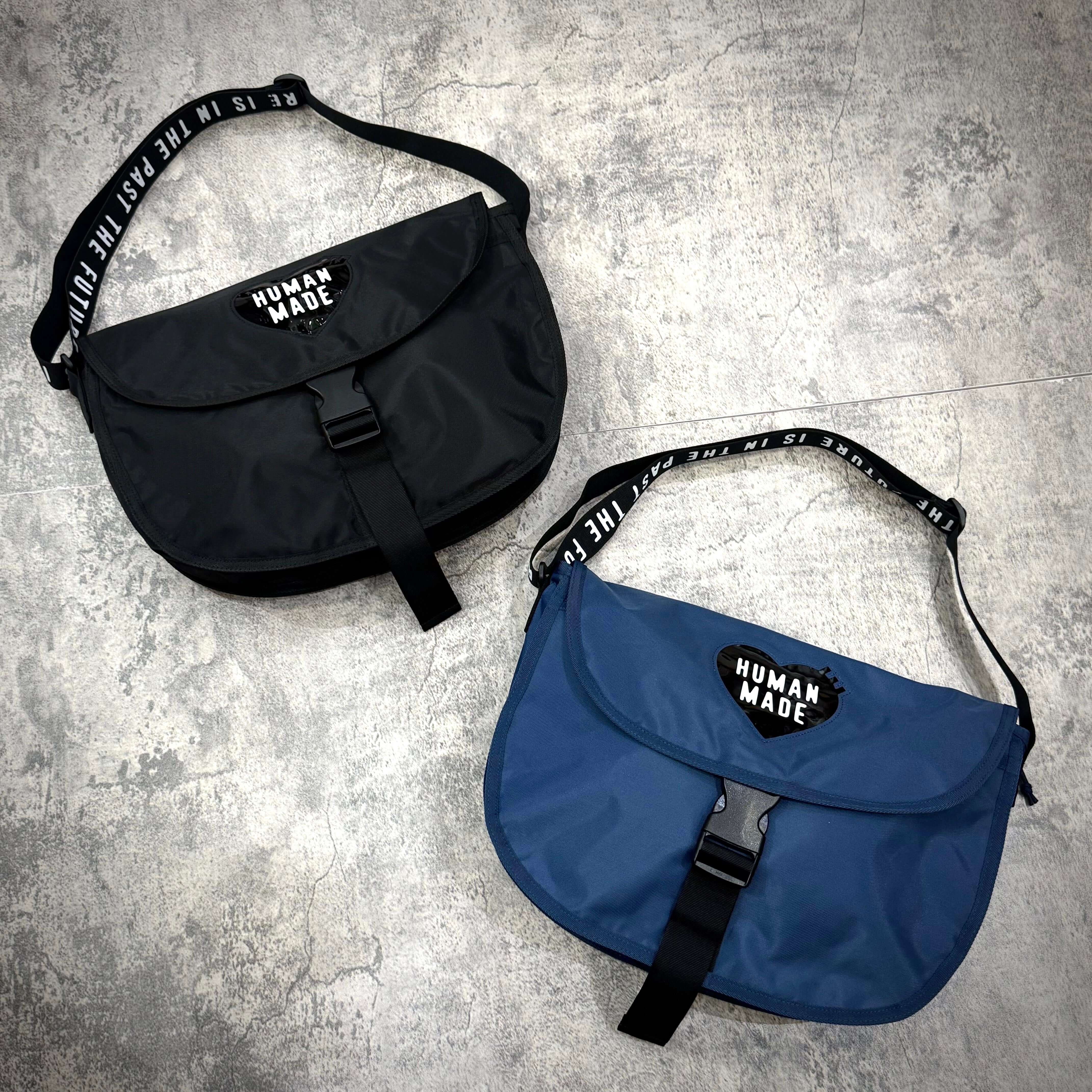 Image of two Human Made Nylon Shoulder Bags, one black and one blue, displayed on a textured surface.