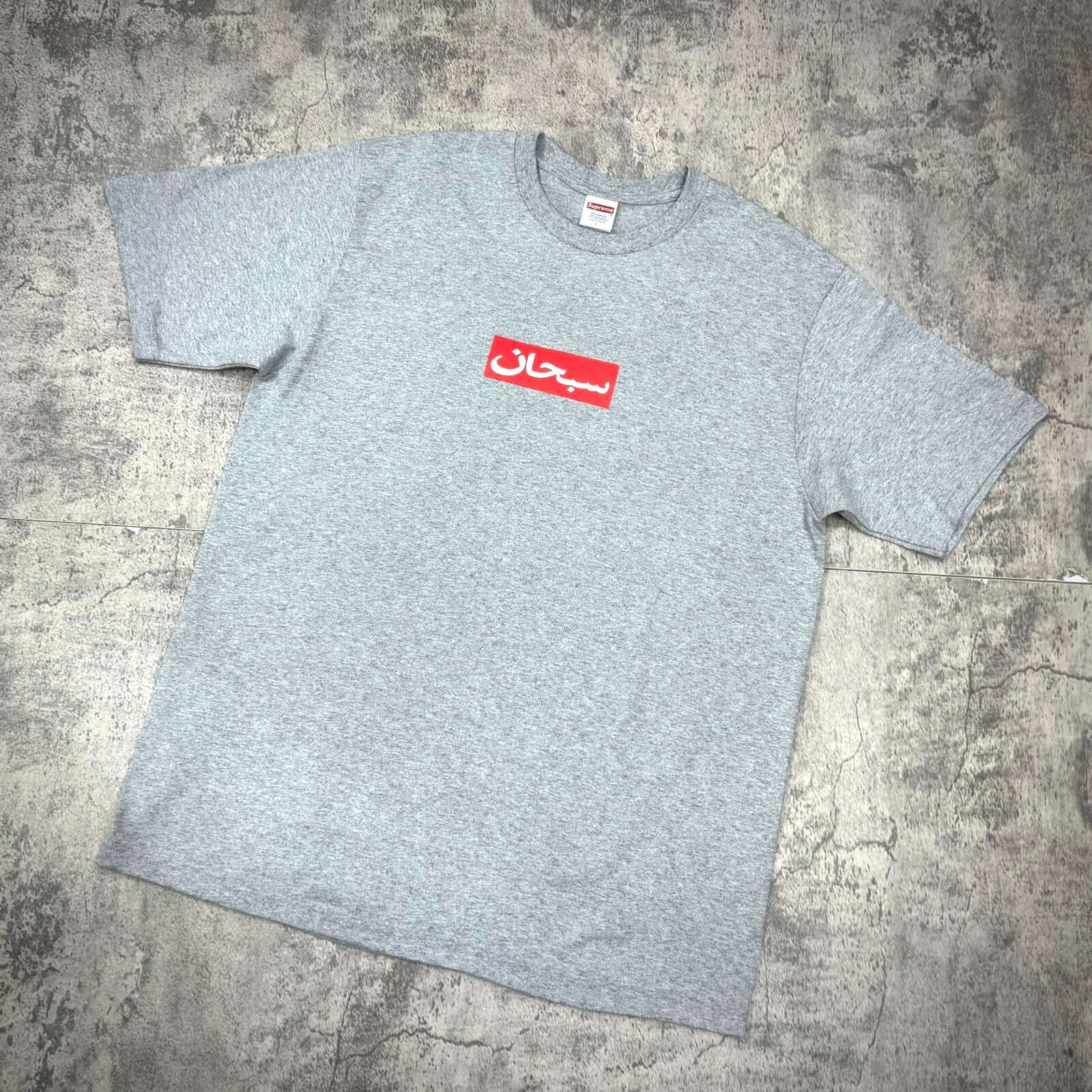 Supreme Arabic Box Logo Tee in gray with red logo, displayed on a textured background.