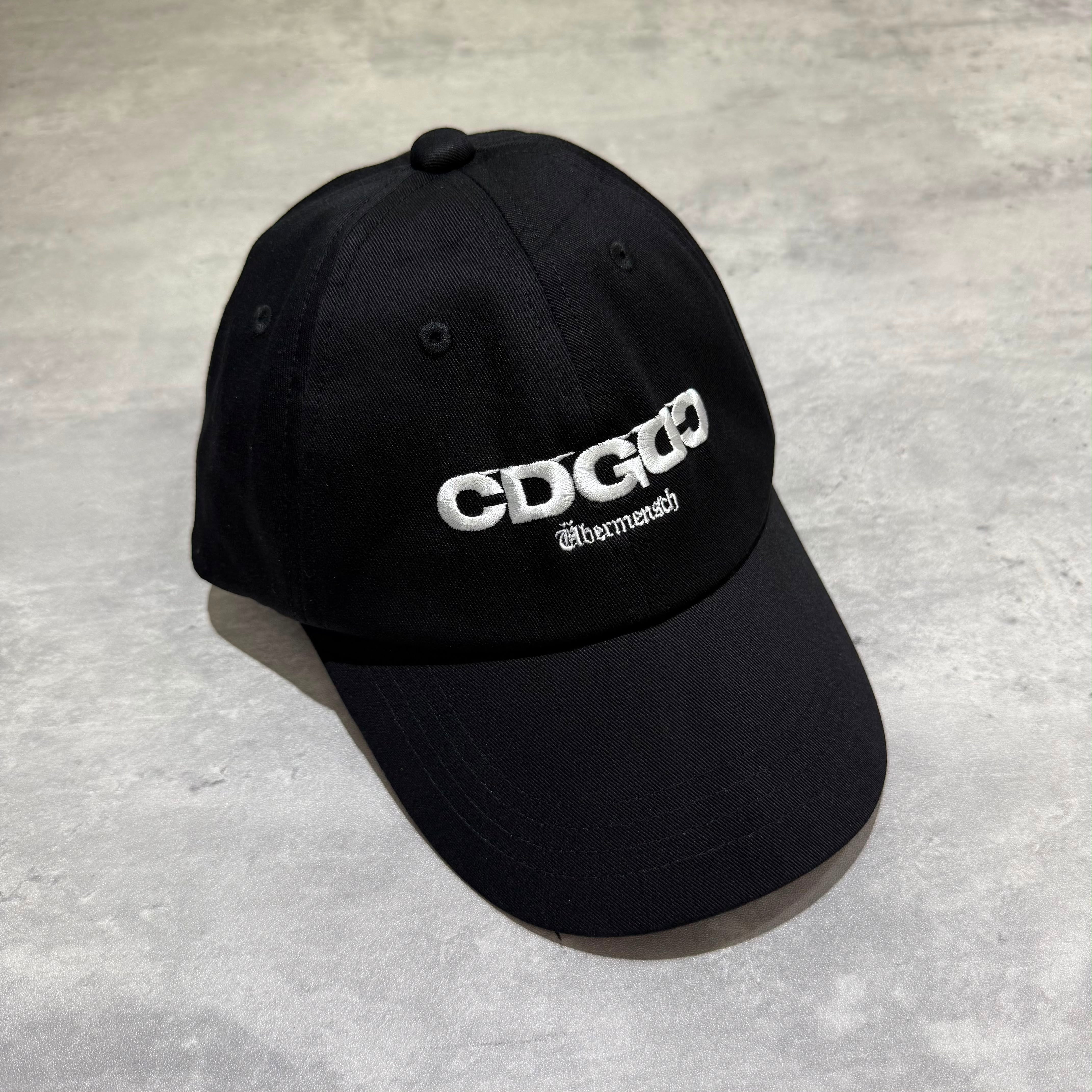 CDG x G-DRAGON Ubermensch Classic Cap displayed on a flat surface with embroidered white logo on black fabric.