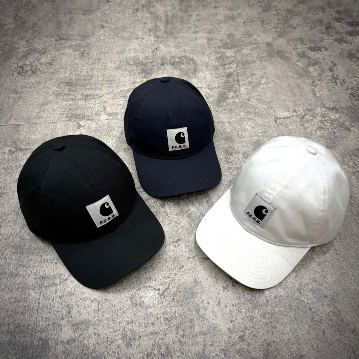 Carhartt WIP x F.C.Real Bristol Cap in black, navy, and white on textured surface.