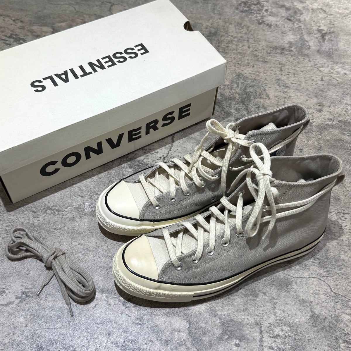 Fear Of God Essentials x Converse Chuck 70s Hi - White / Grey sneakers next to the box on a textured surface.