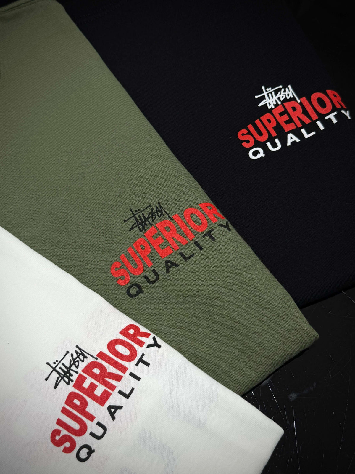 Stussy Superior Quality Tee - Black, green, and white shirts displaying the Superior Quality logo on a dark background.