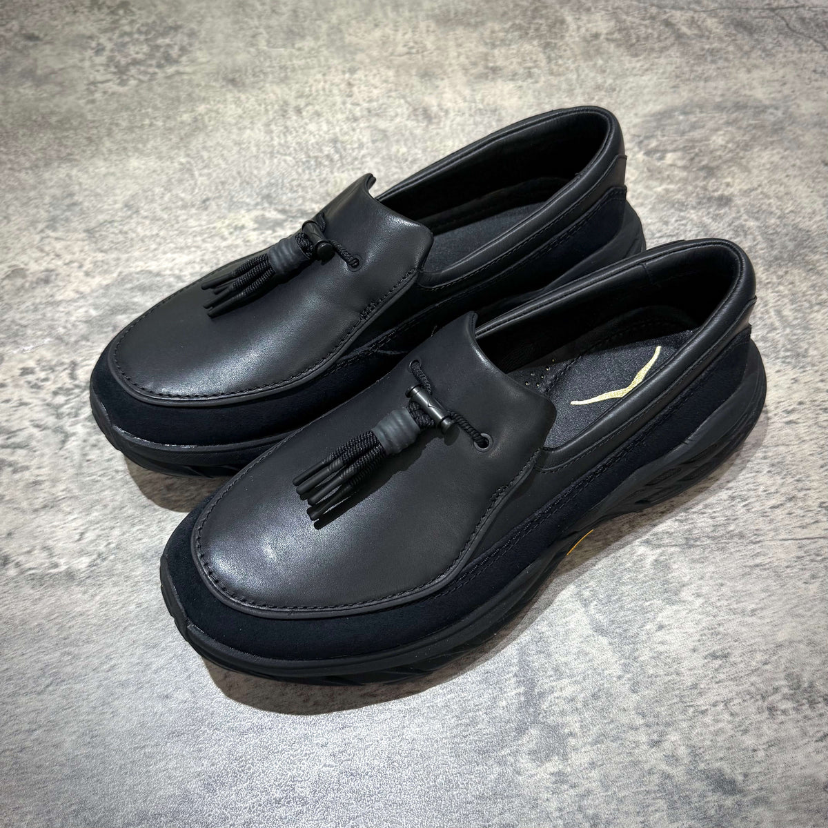 Hoka Speed Loafers - Black 1162170-BBLC displayed on a textured surface, showcasing their sleek design and unique tassel detail.