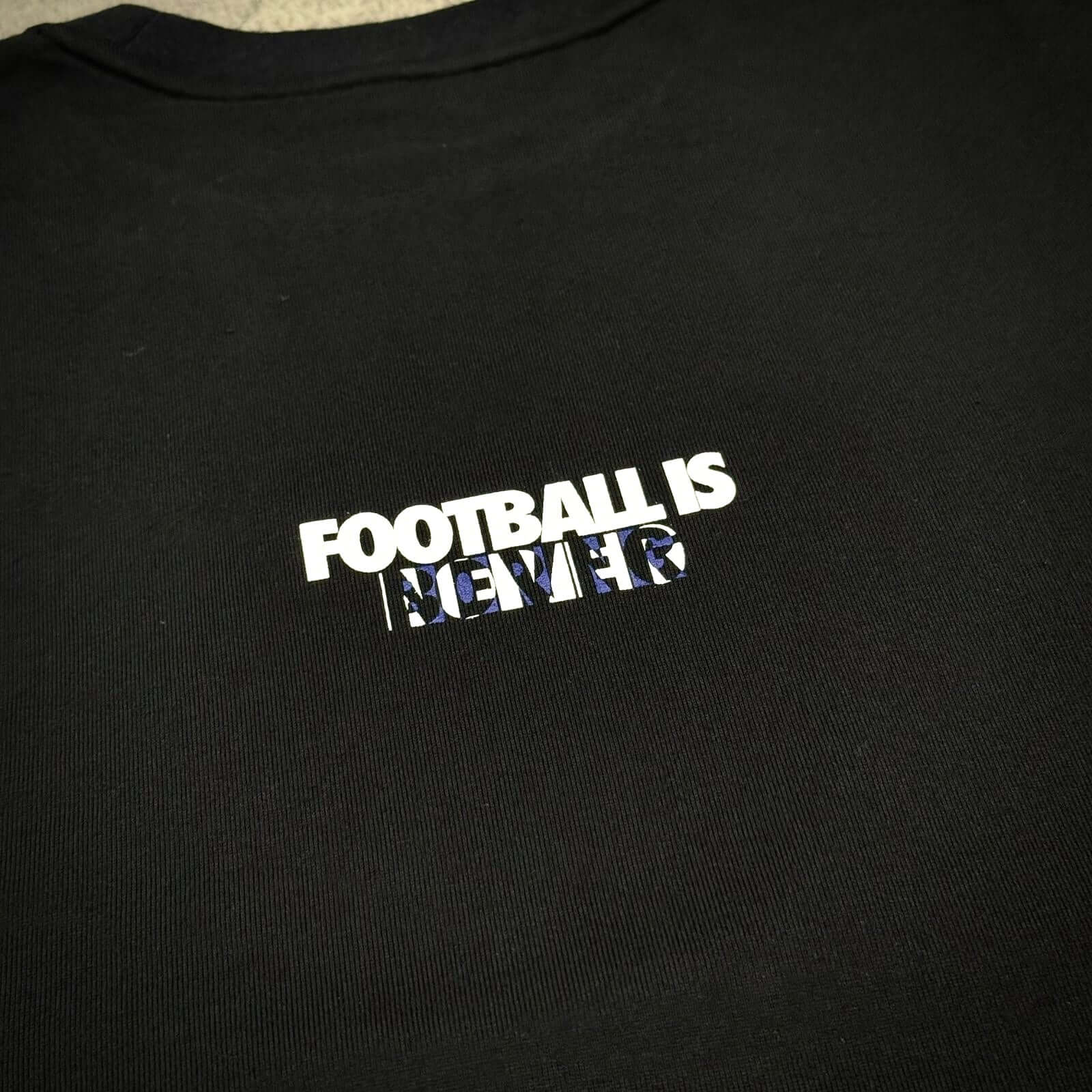 Close-up of the Kojiro Hyuga x FCRB Emblem Tee featuring the text 'FOOTBALL IS NEVER' in white on a black background.