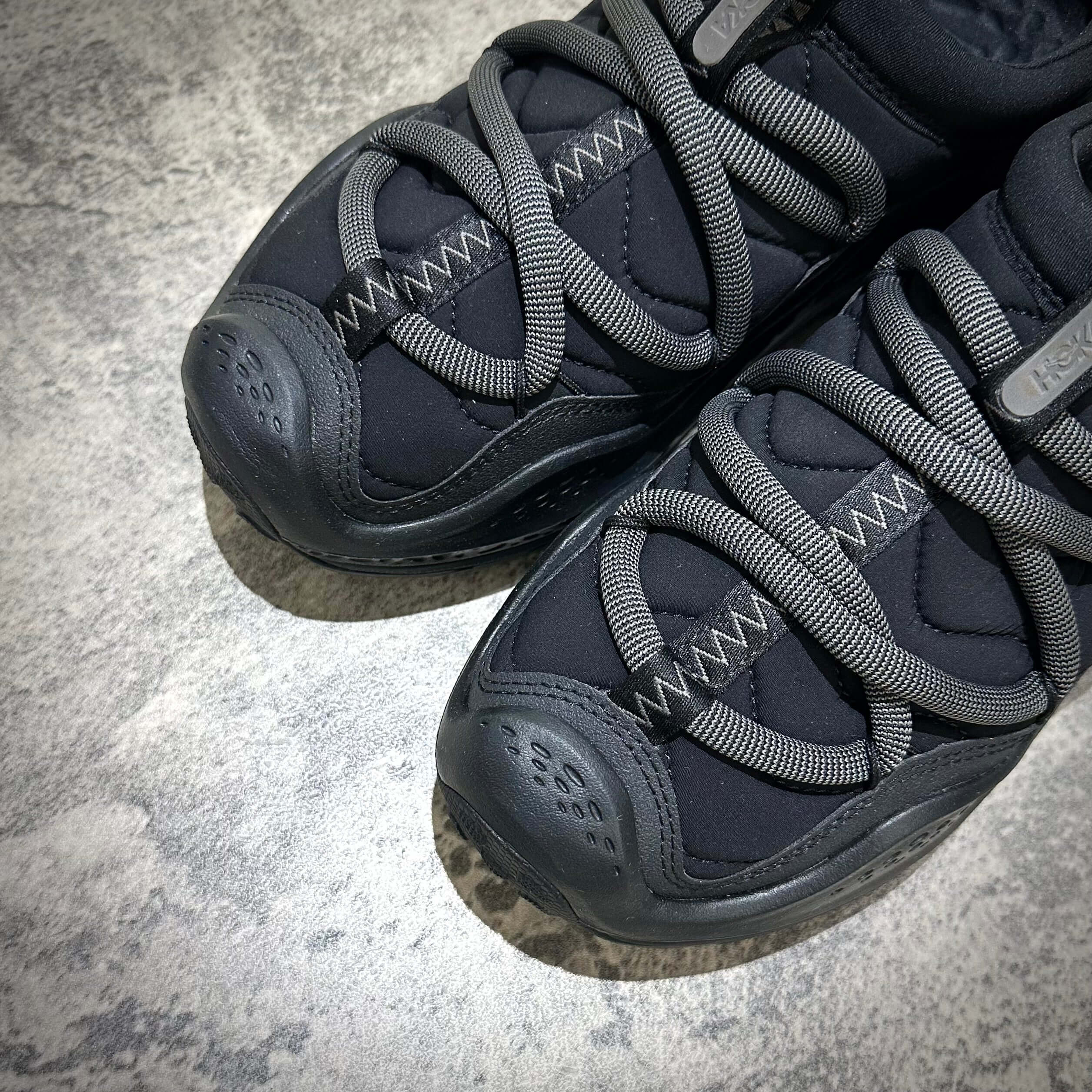 Hoka Ora Primo - Black sneaker features close-up view with gray laces on a textured surface.