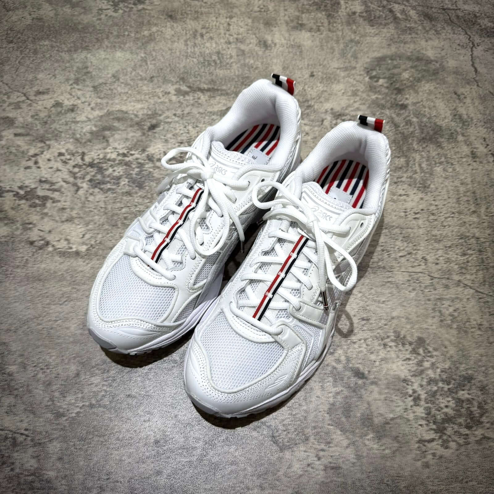 Thom Browne x Asics Gel-Kayano 14 - White sneakers with red and blue accents on a textured surface.