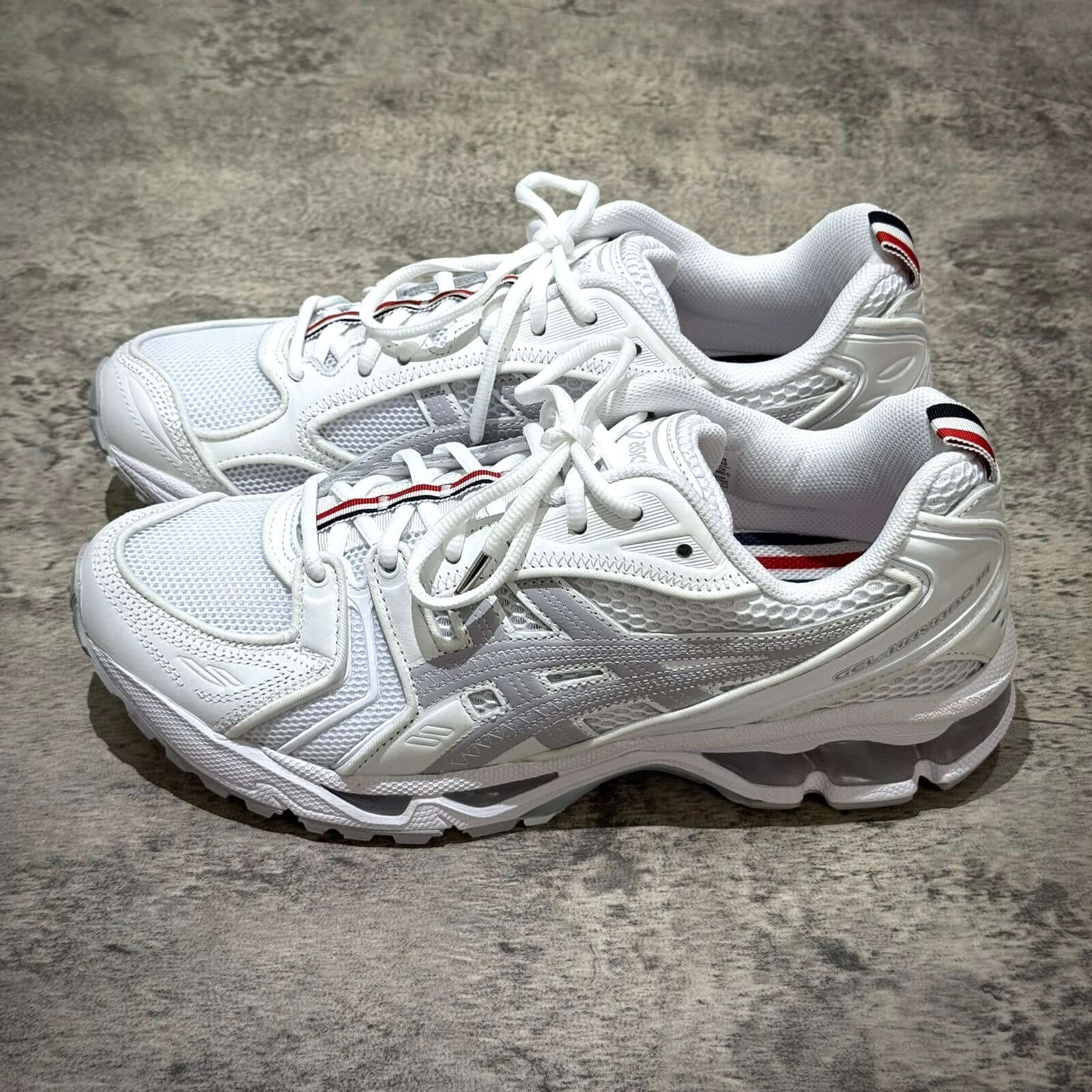 Thom Browne x Asics Gel-Kayano 14 - White sneakers showing sleek design and signature colors on a textured floor.