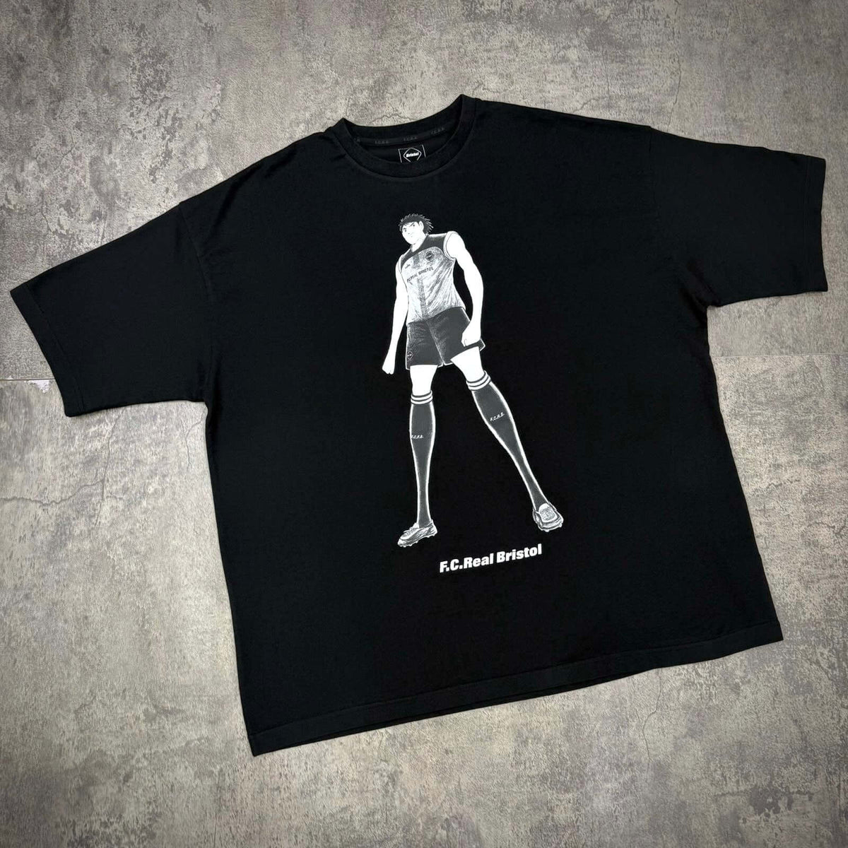 Kojiro Hyuga x F.C.Real Bristol On The Front Baggy Tee featuring a graphic of a soccer player in black.