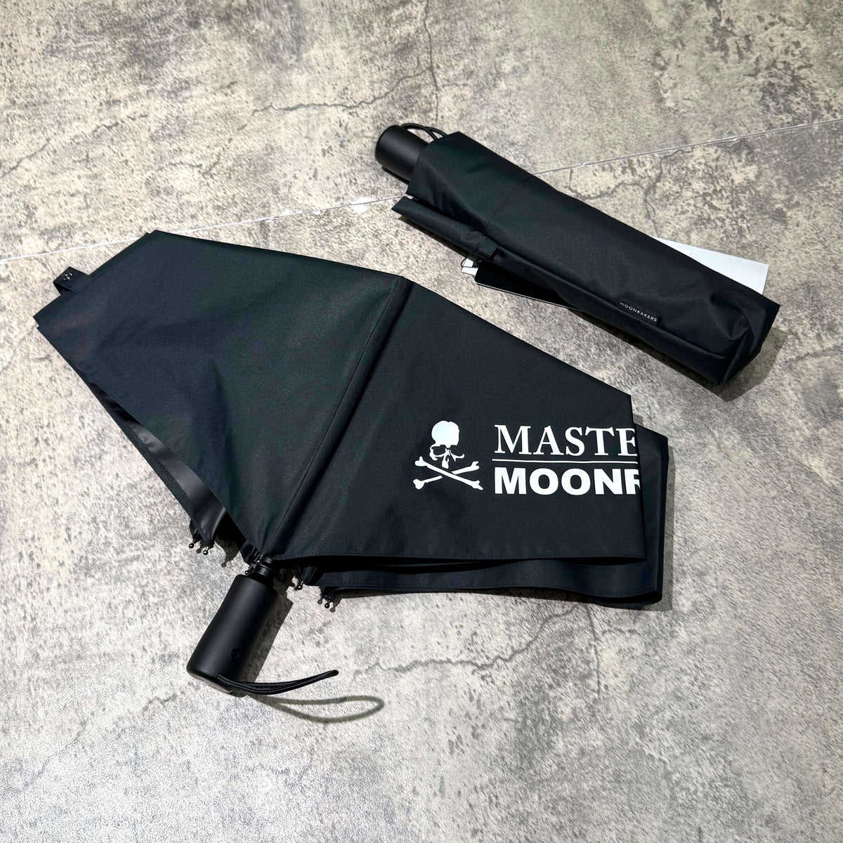 Mastermind Japan Moonrakers Umbrella in black with logo and compact case on a textured surface.