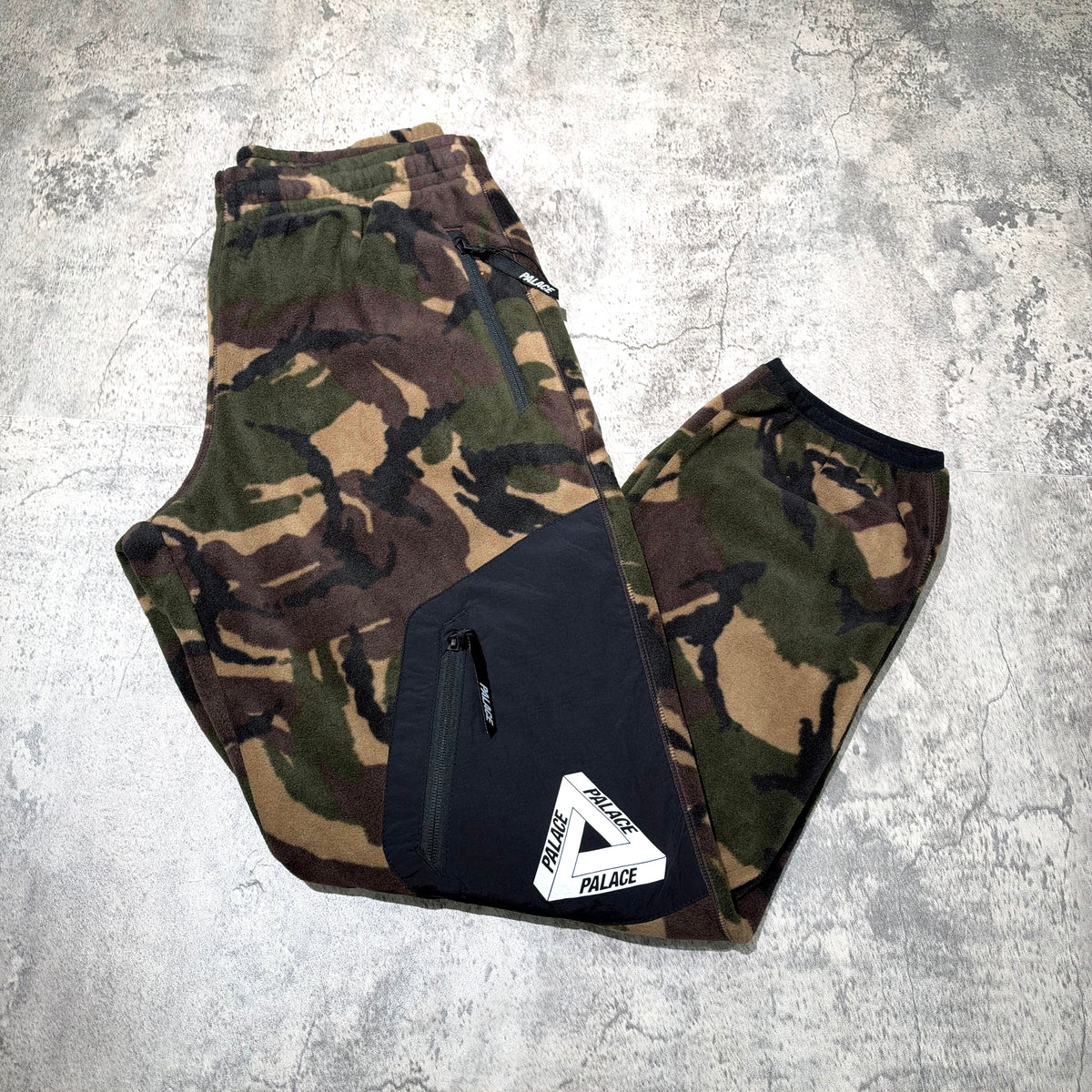 Palace P-Surgent Fleece Joggers Black DPM displayed on a textured surface, featuring camouflage pattern and zip pocket.