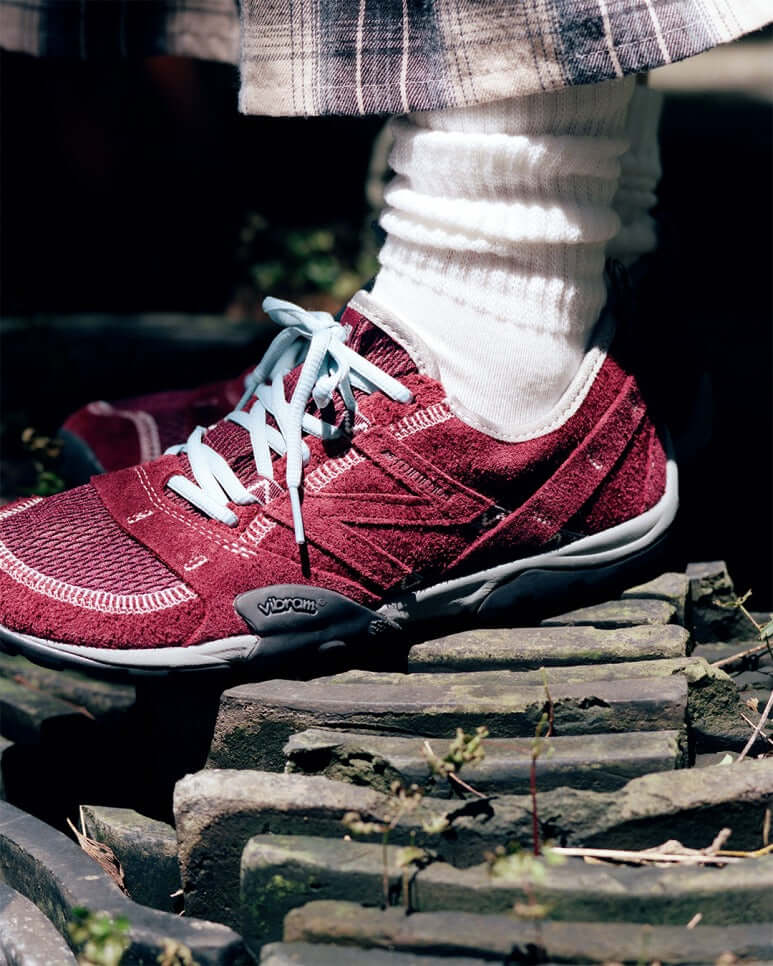 Model wearing SUNLOVETOUR x New Balance MT1001ND shoes in burgundy on rocky terrain.