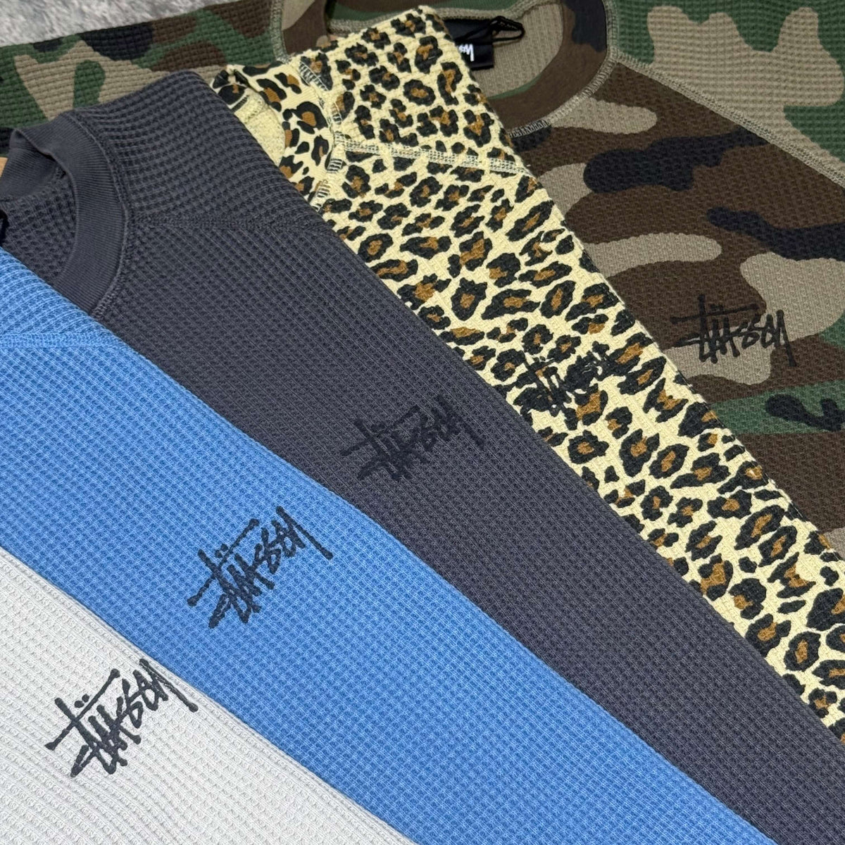 Stussy Basic Raglan Thermal LS Crew FW26 collection featuring various colors and prints laid out on a surface.