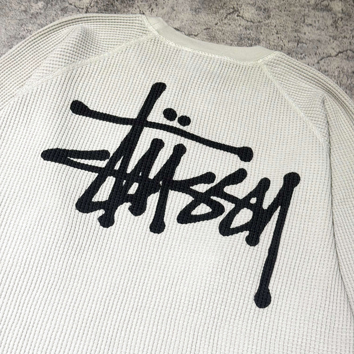 Stussy Basic Raglan Thermal LS Crew FW26 featuring iconic logo on the back with a textured white backdrop.