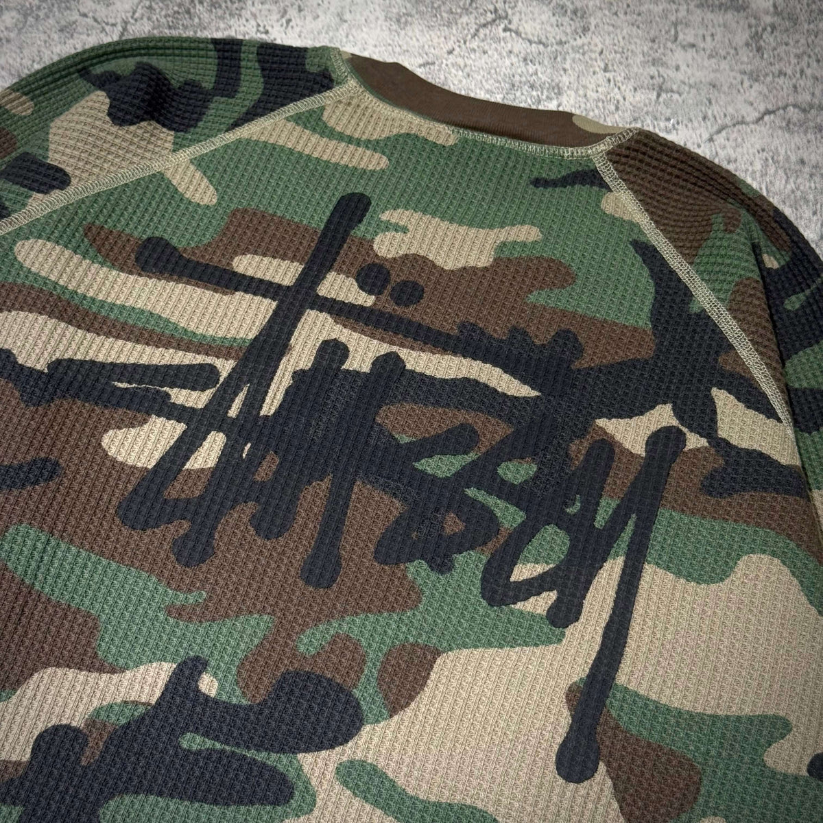 Stussy Basic Raglan Thermal LS Crew FW26 in camo print with bold black logo design on textured fabric.