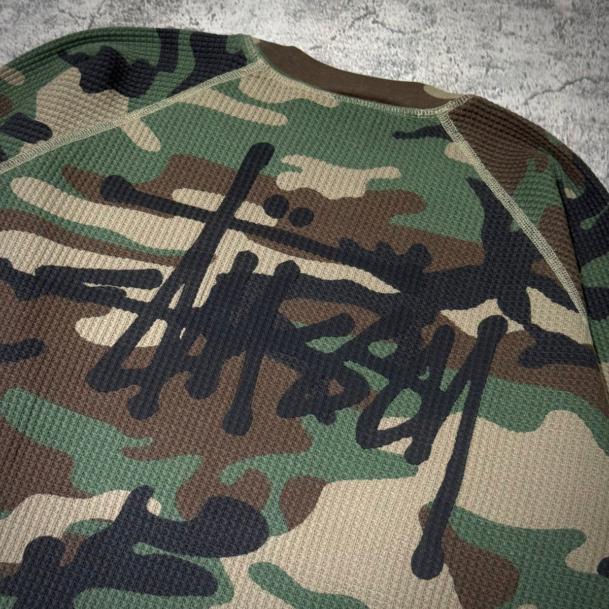 Stussy Basic Raglan Thermal LS Crew FW26 in camo print with bold black logo design on textured fabric.