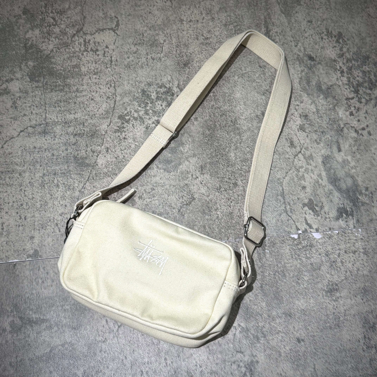 Stussy Canvas Side Pouch on a textured surface, showcasing a neutral color and adjustable strap.