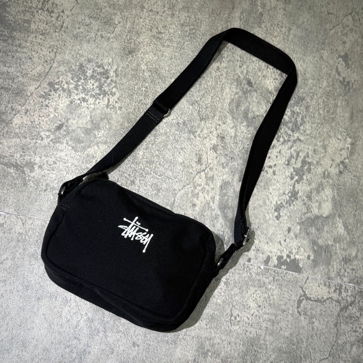 Stussy Canvas Side Pouch in black with adjustable strap on a textured surface.