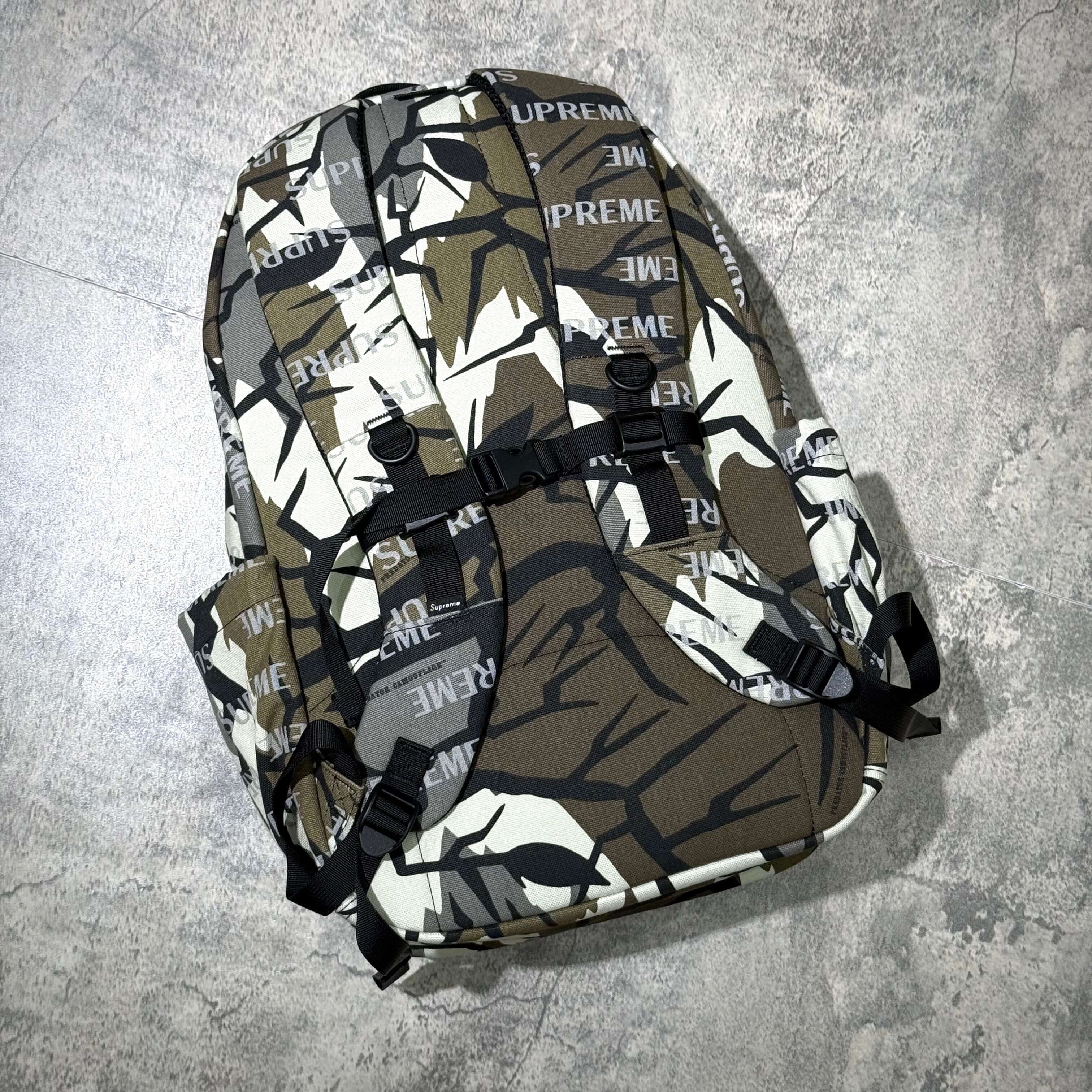 Supreme FW26 Backpack featuring a camo pattern with bold SUPREME branding and adjustable straps on a textured surface.