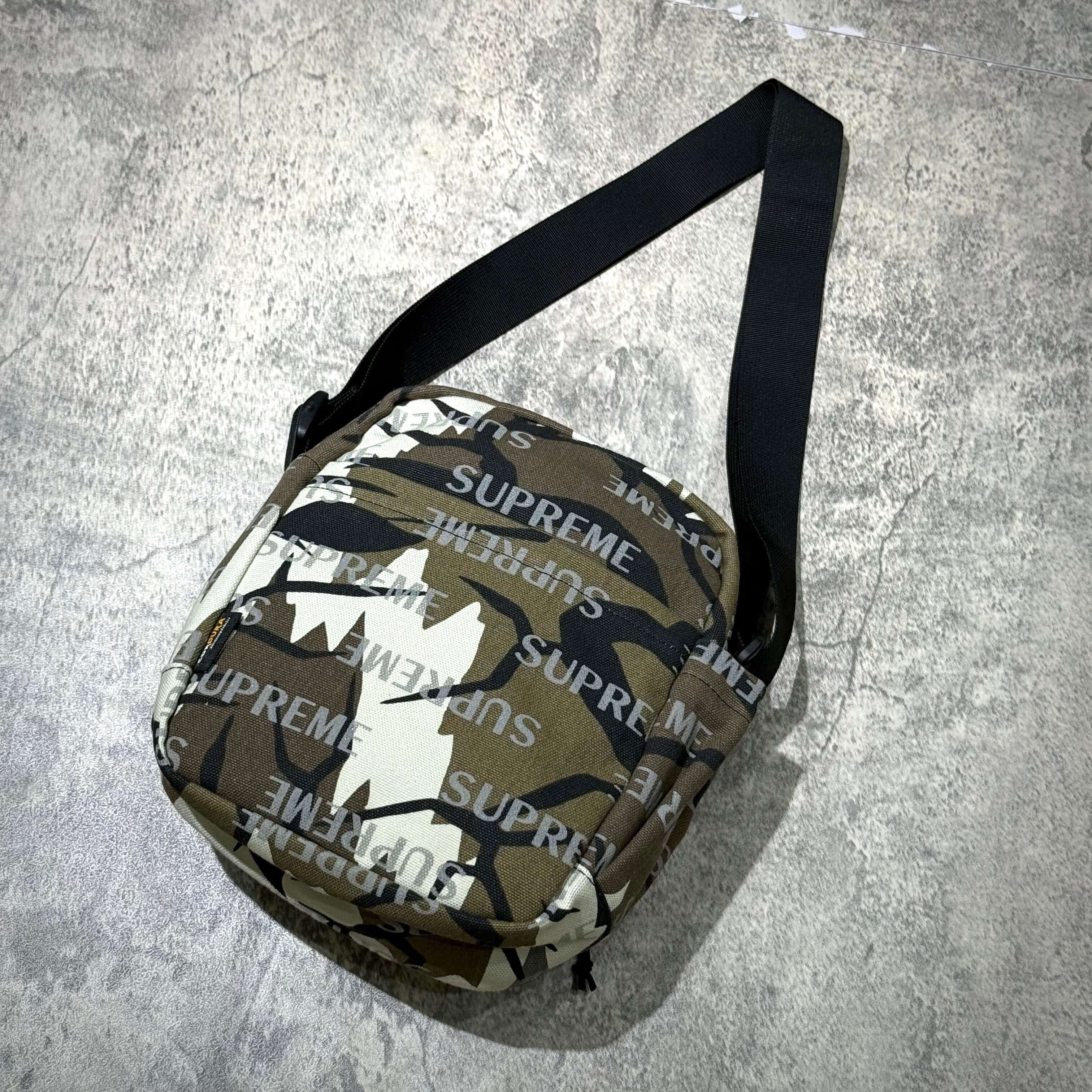 Supreme FW26 Shoulder Bag featuring a camo design and adjustable strap on a textured surface.