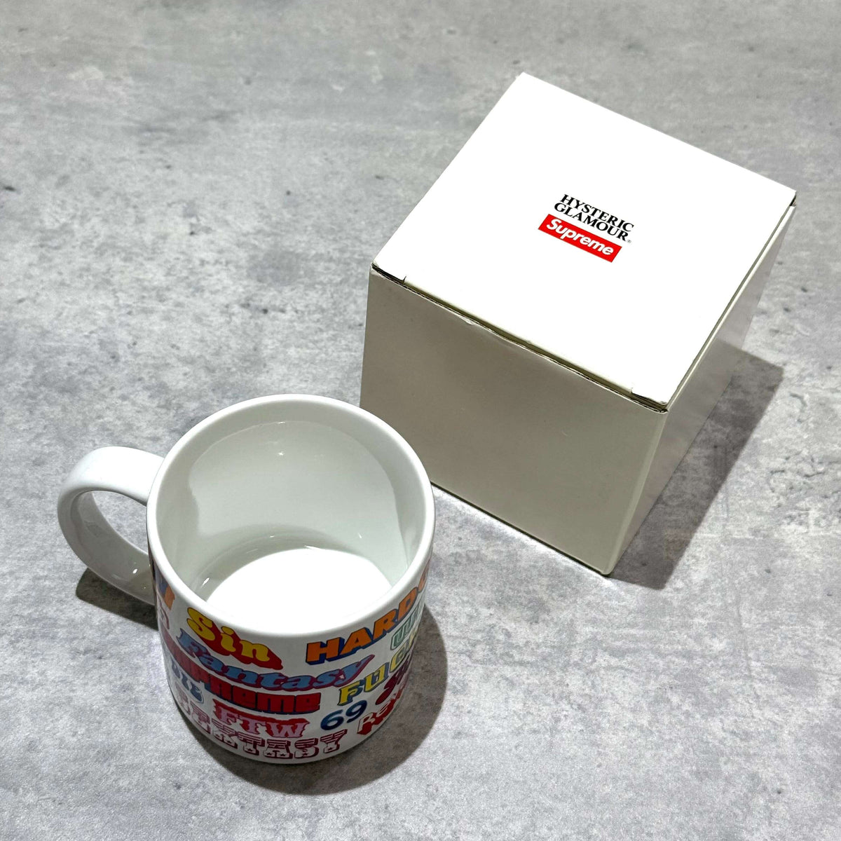Supreme Hysteric Glamour Ceramic Coffee Mug displayed next to its box on a textured surface.