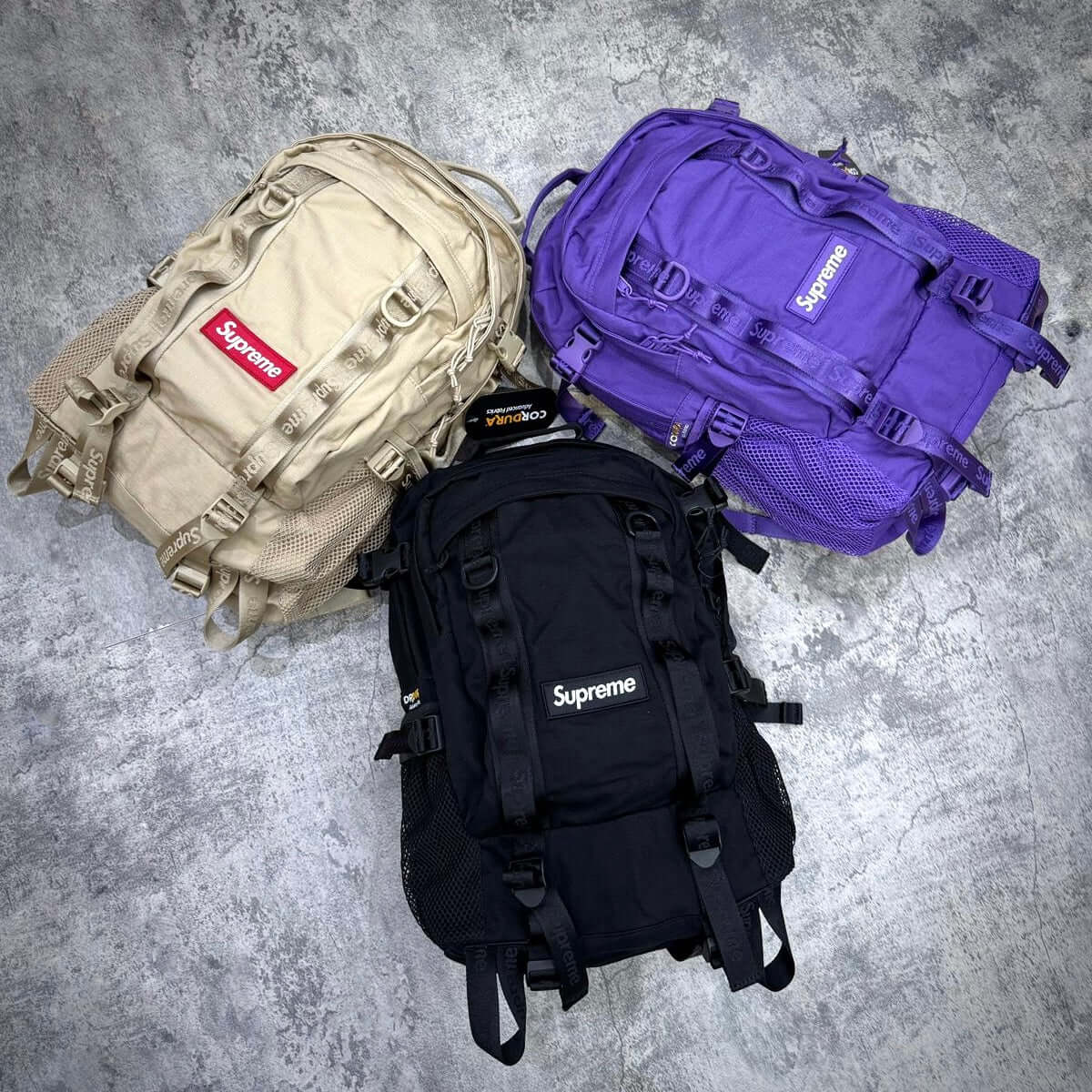 Supreme Backpack SS26 in black, purple, and beige color variations displayed on a textured surface.