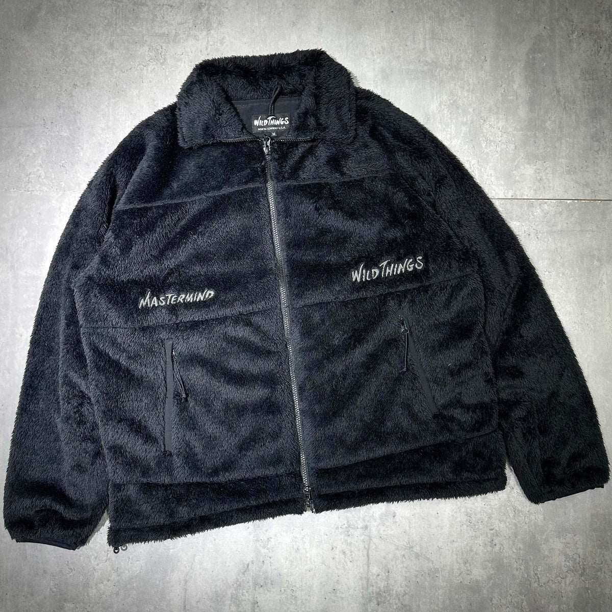 Wild Things x Mastermind World Shaggy Zip Up Jacket displayed on a gray background, showcasing its cozy black fleece material.