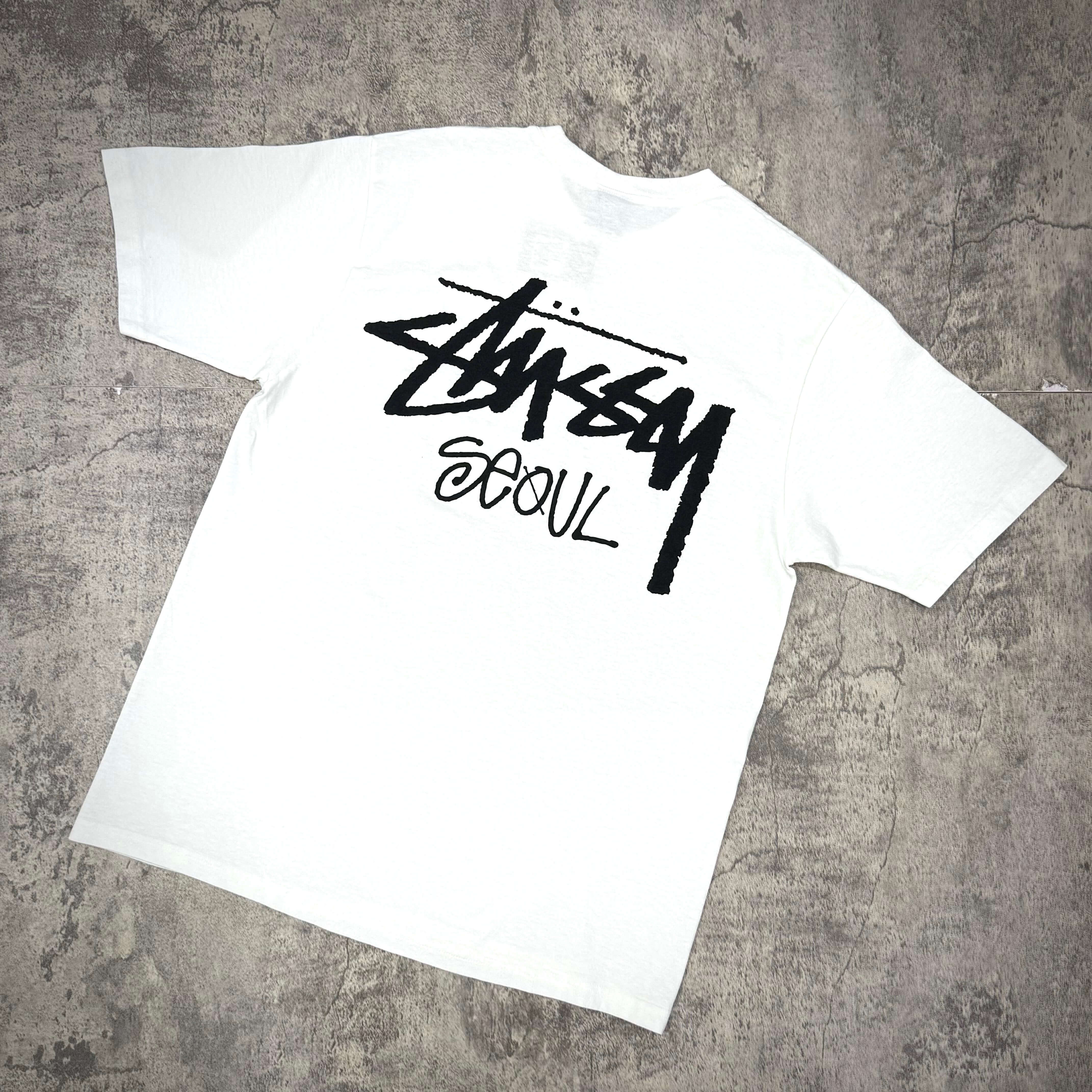 Stussy Seoul Exclusive Logo Tee in white with bold black logo on a textured background.