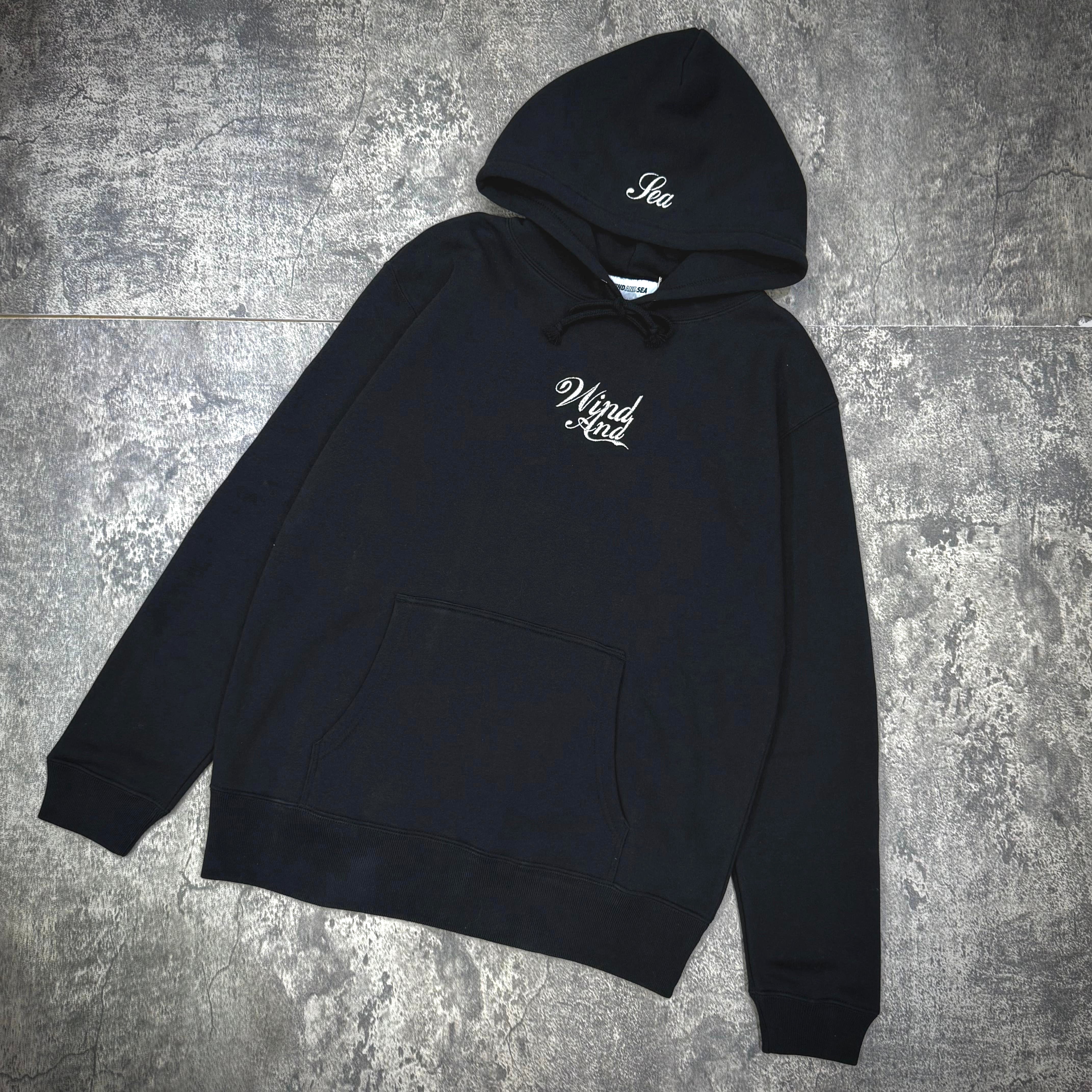 Wind And Sea Glitter Hoodie - Black / White displayed on a textured surface with a front pocket and logo.