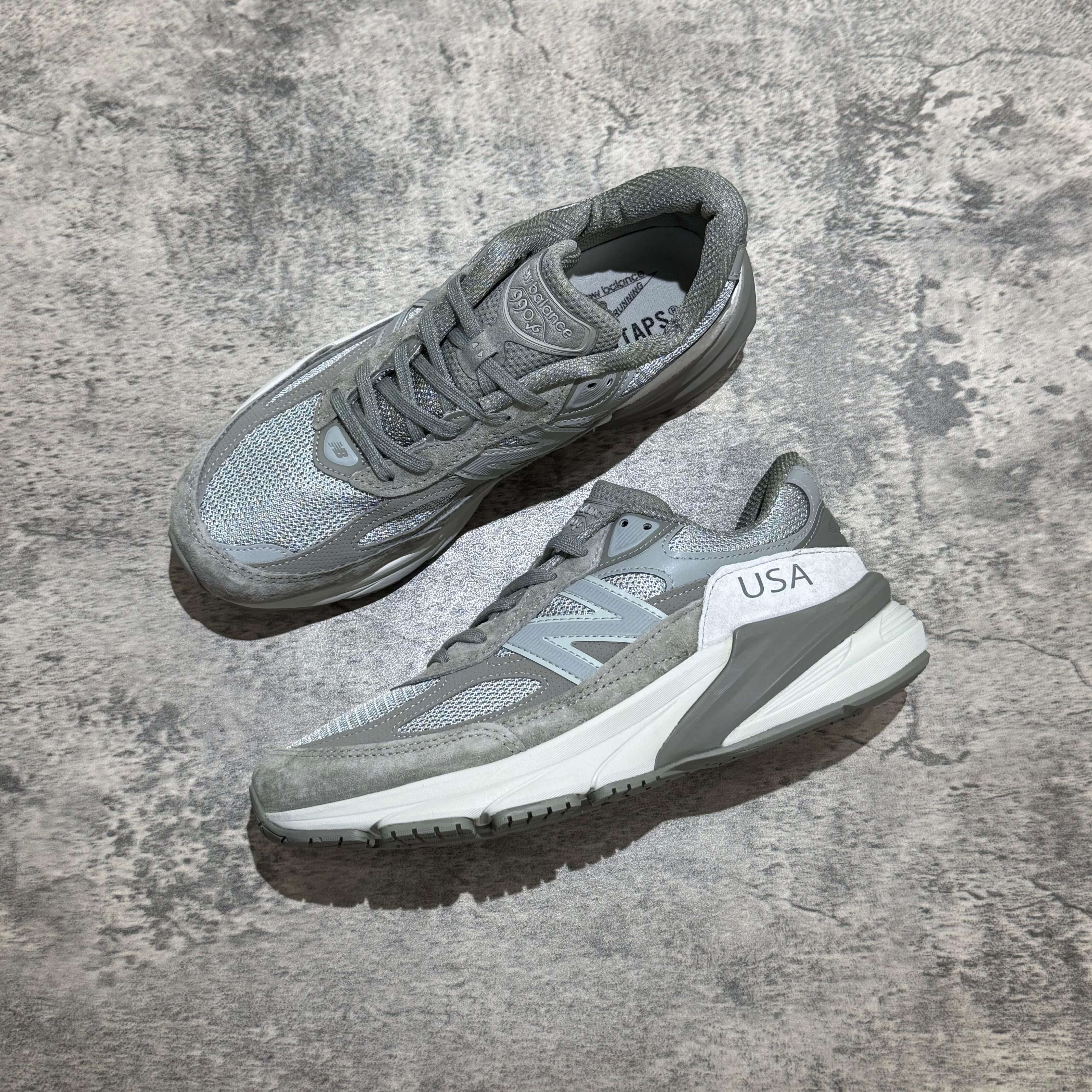 New Balance x Wtaps 990v6 grey sneakers on a textured surface, showcasing a stylish athletic design.