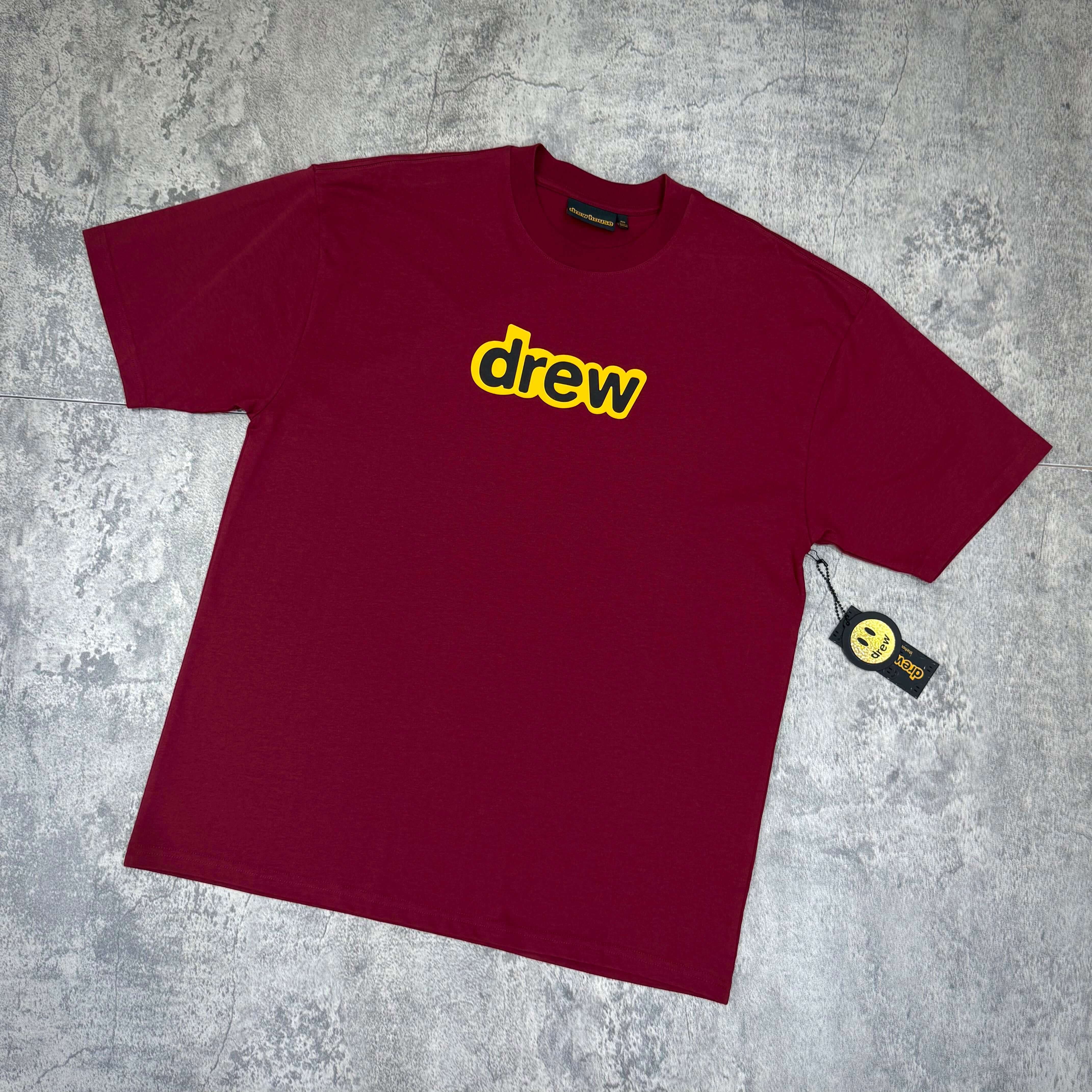 Drew House Secret SS Tee - Burgundy displayed on a gray background, featuring a yellow "drew" logo.