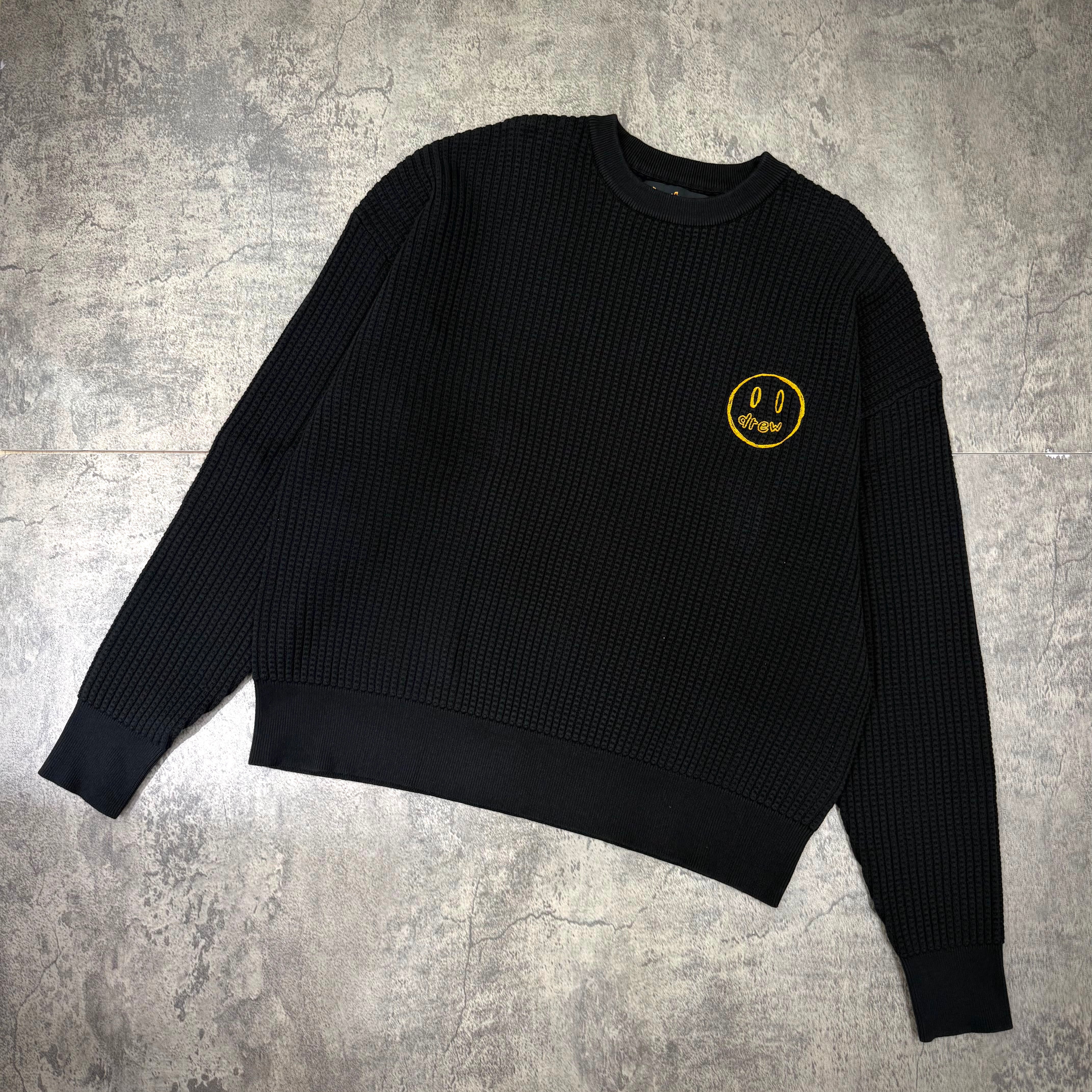 Drew House Mascot Waffle Sweater - Black with yellow smiley logo on the chest, displayed flat on a textured surface.