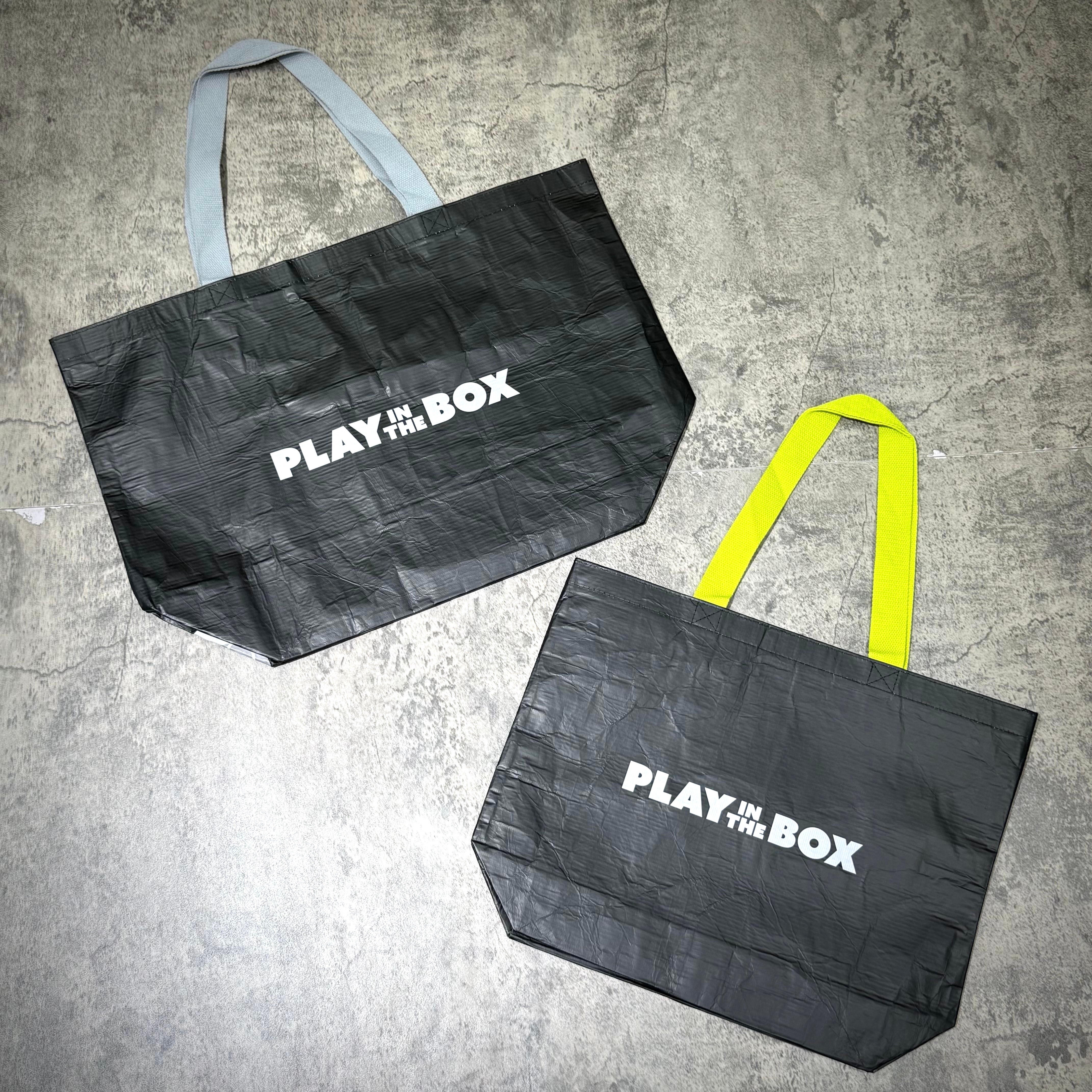 Two reusable bags with the text 'PLAY IN THE BOX' displayed, one with gray handles and the other with yellow handles.