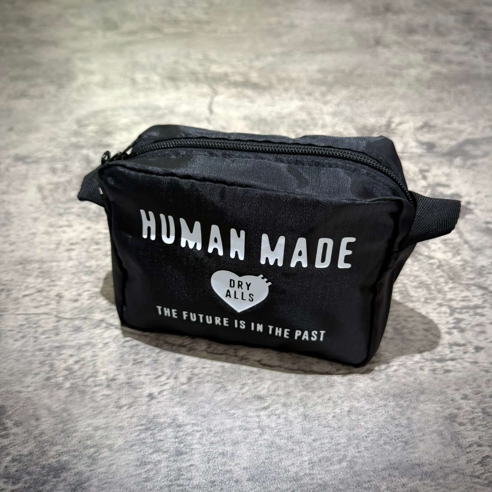 Human Made Travel Pouch displayed on a textured surface, featuring a black design and unique text.