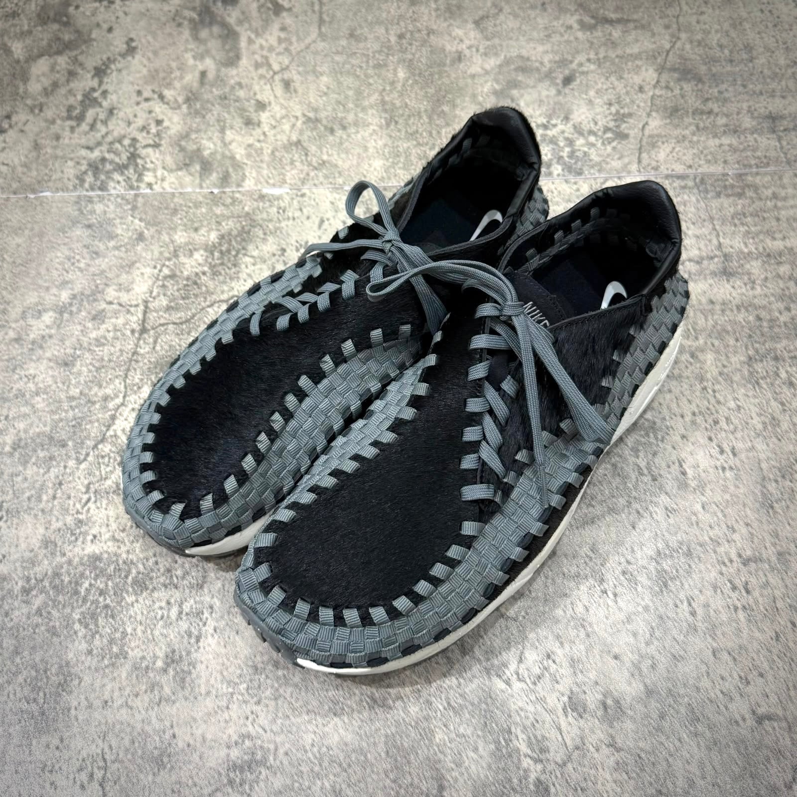Nike Air Footscape Woven Black Smoke Grey shoes with woven design and laces on a textured surface.
