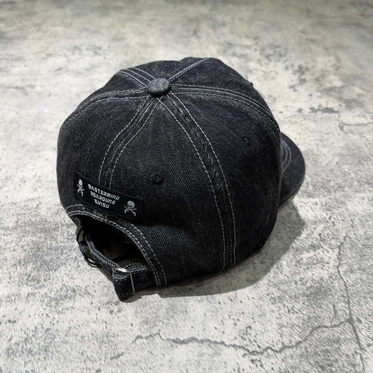 Evisu x Mastermind Japan x Roarguns Cap, black denim cap with logo tag, back view on a textured surface.
