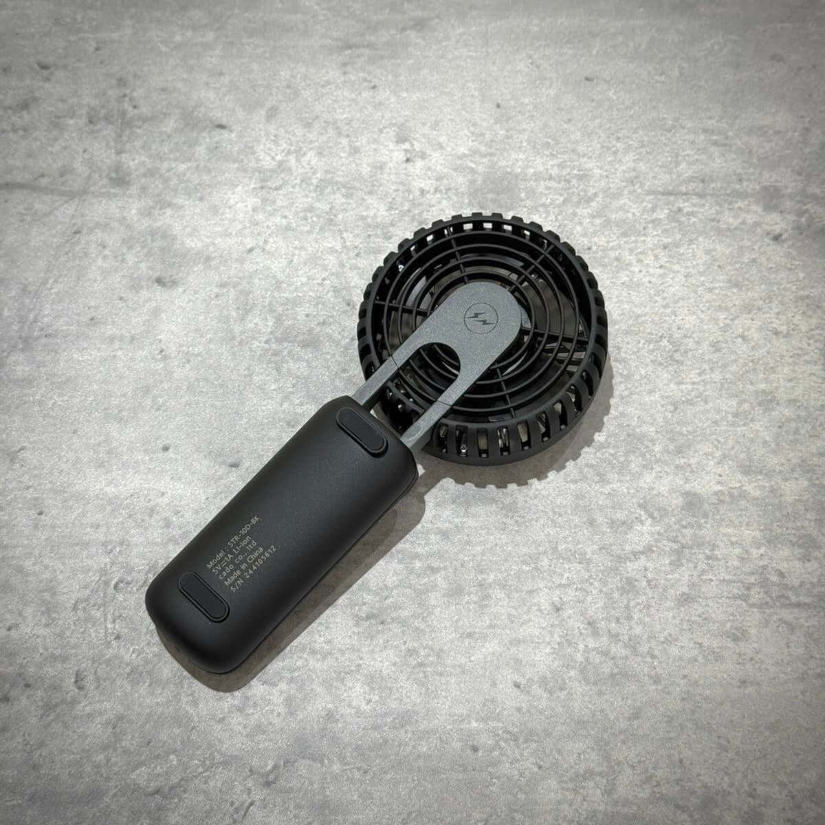 Fragment Design x Cado Mini Handy Fan on a textured surface, showcasing its sleek black design and compact size.