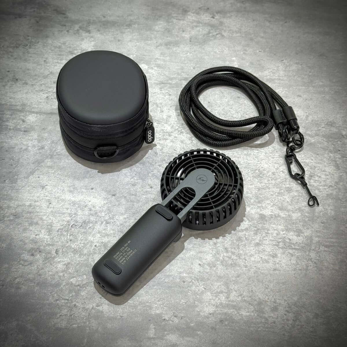 Fragment Design x Cado Mini Handy Fan with carrying case and strap accessories on a textured surface.