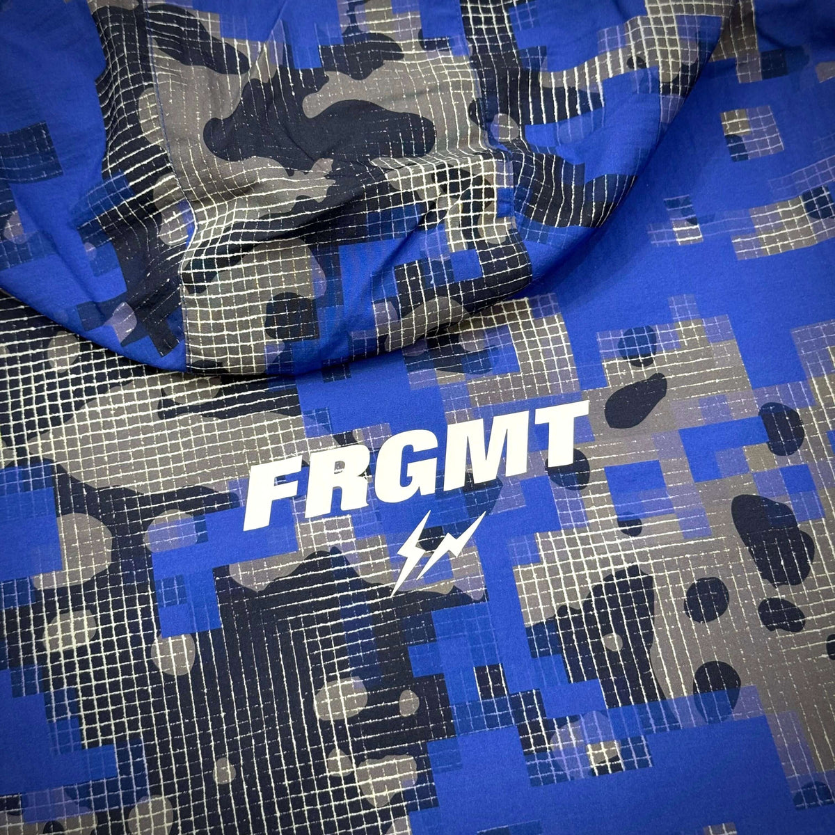 Fragment x F.C.R.B. Camouflage Nylon Blouson的藍色迷彩設計，展示FRGMT字樣，展現時尚潮流風格。
