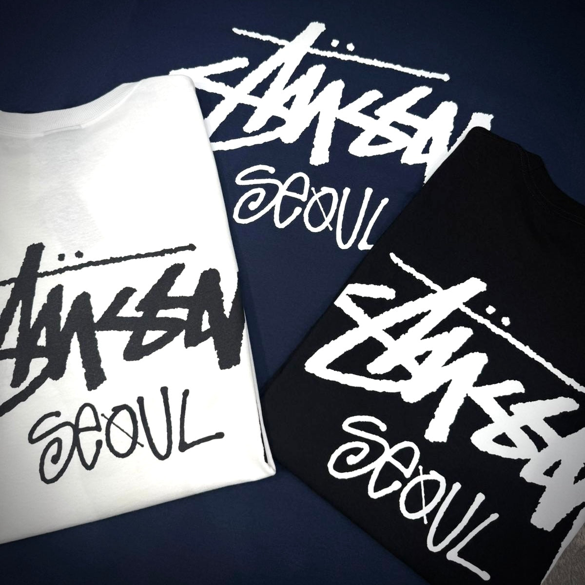 Stussy Seoul Exclusive Logo Tee in black and white displayed on a dark background.