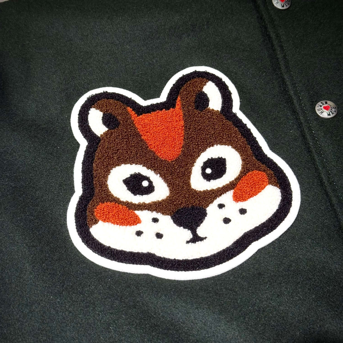Image of a cute embroidered animal patch featuring a stylized chipmunk face on a dark green fabric.