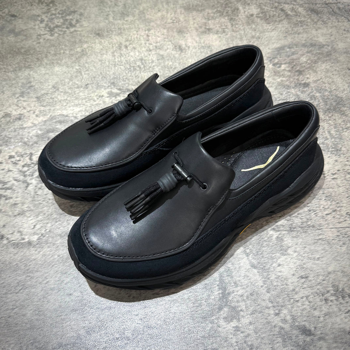 Hoka Speed Loafers - Black 1162170-BBLC displayed on a textured surface, showcasing their sleek design and unique tassel detail.