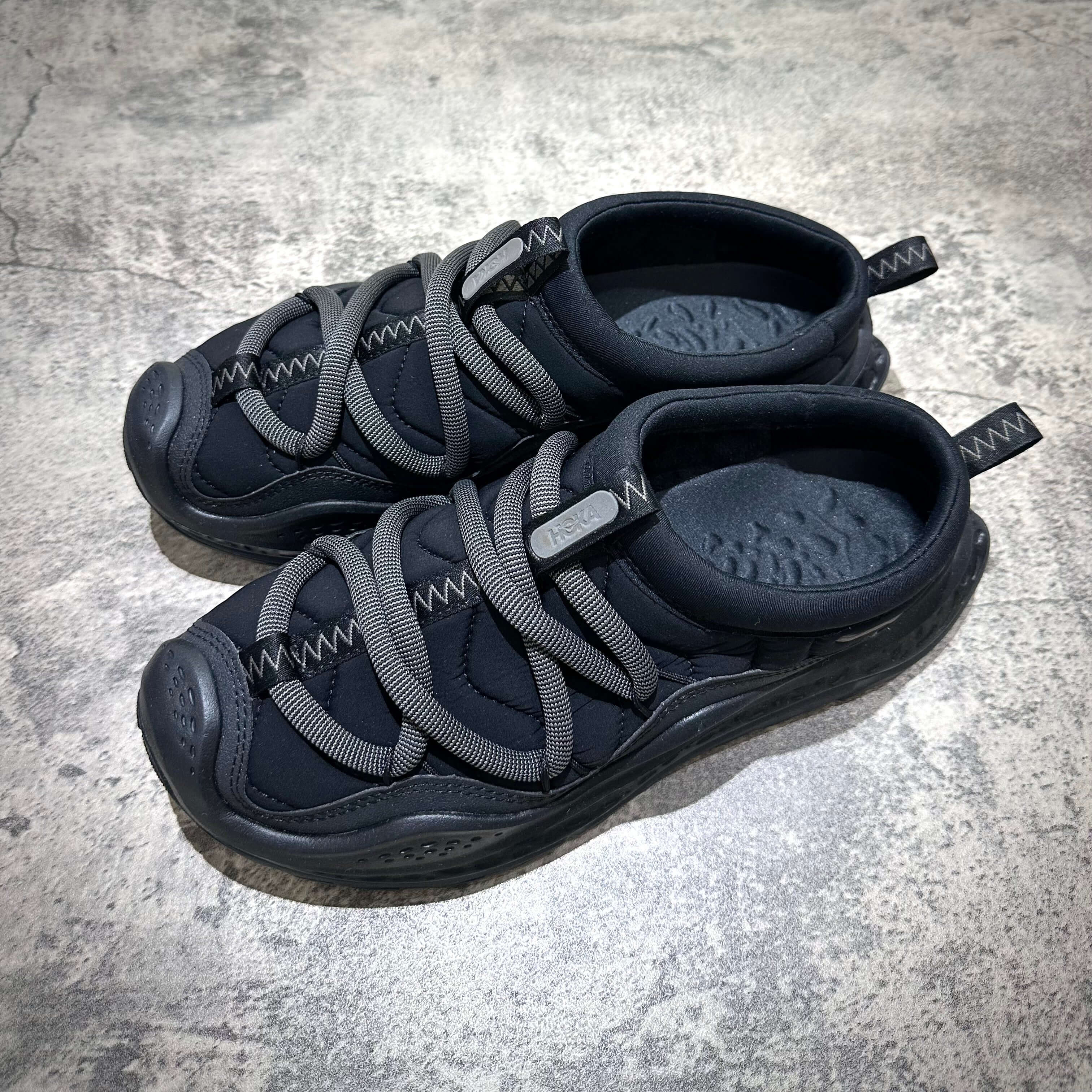 Hoka Ora Primo - Black shoes with unique gray laces on a textured surface, showcasing a modern athletic design.