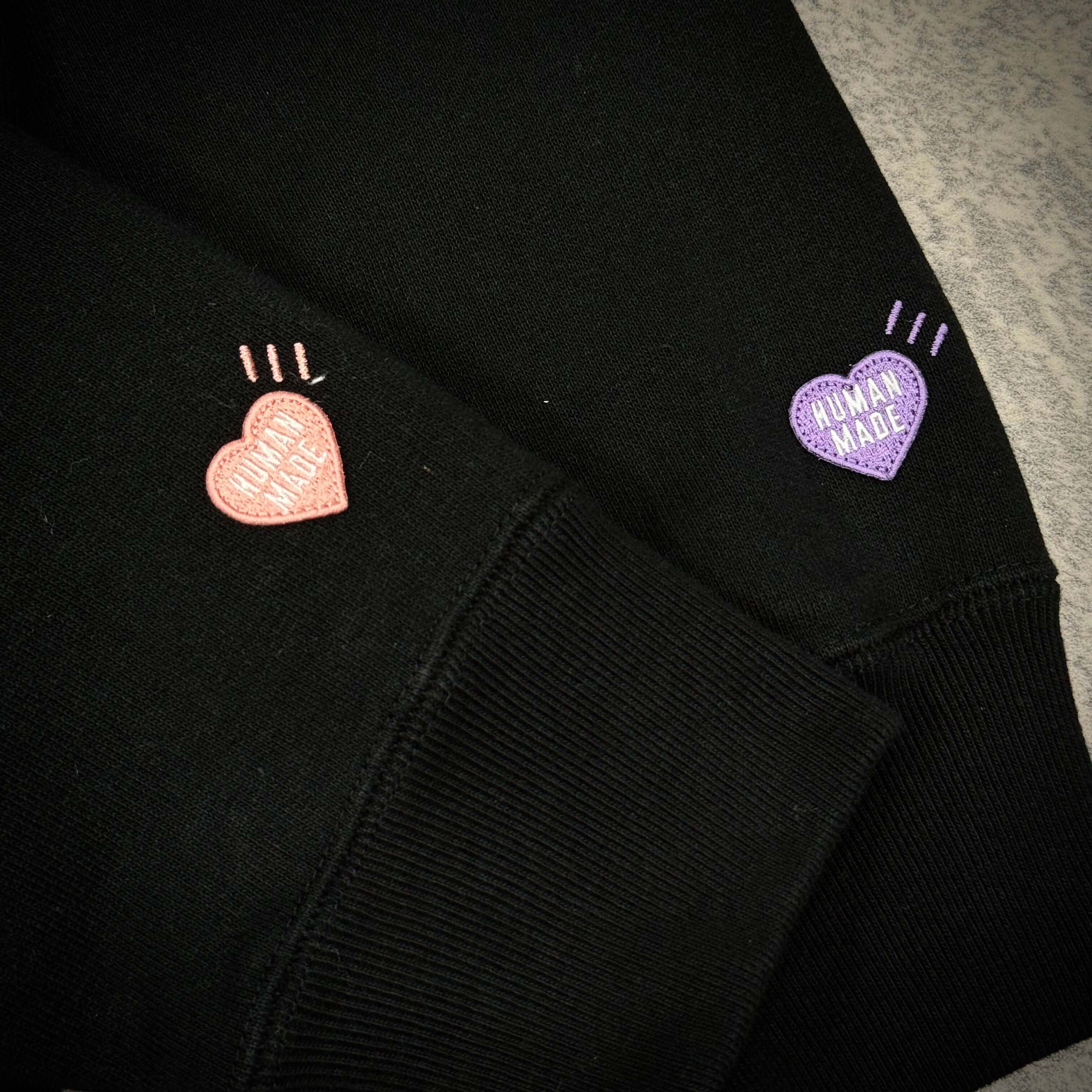 Close-up of black hoodie sleeves featuring pink and purple Human Made Korea heart logos.