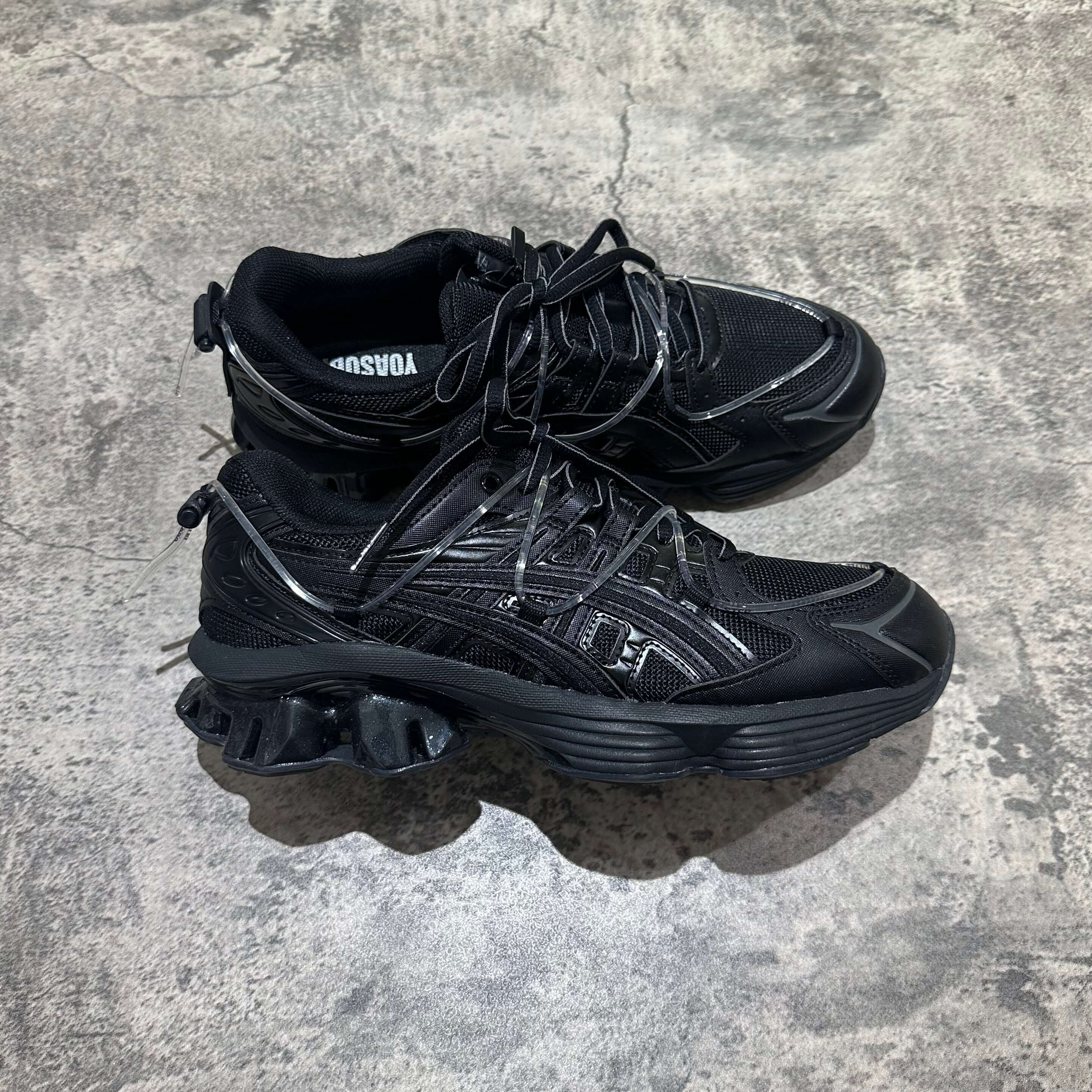 Yoasobi x Asics Gel Kinetic Fluent Y - Black sneakers on a textured surface, showcasing the sleek black design and details.
