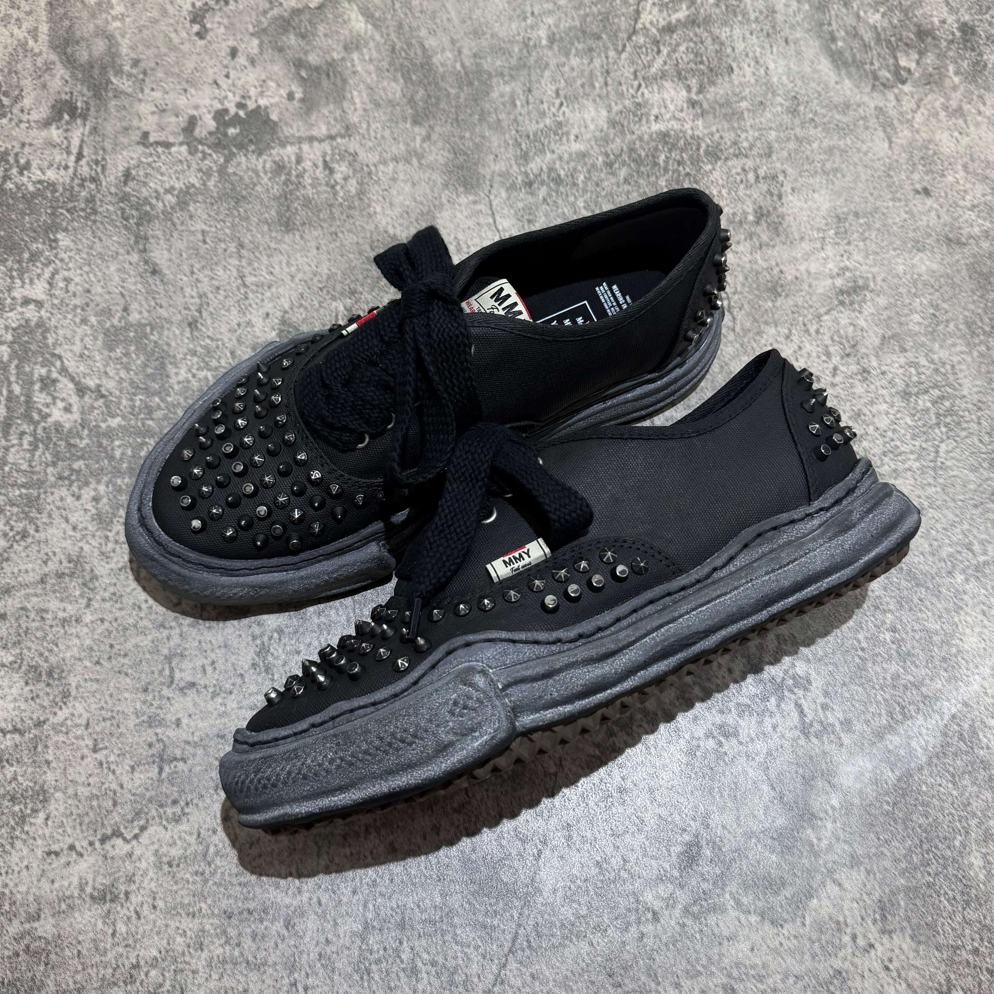 MMY Baker Studded Canvas Low - All Black shoes with studded design and black laces on a textured gray surface.
