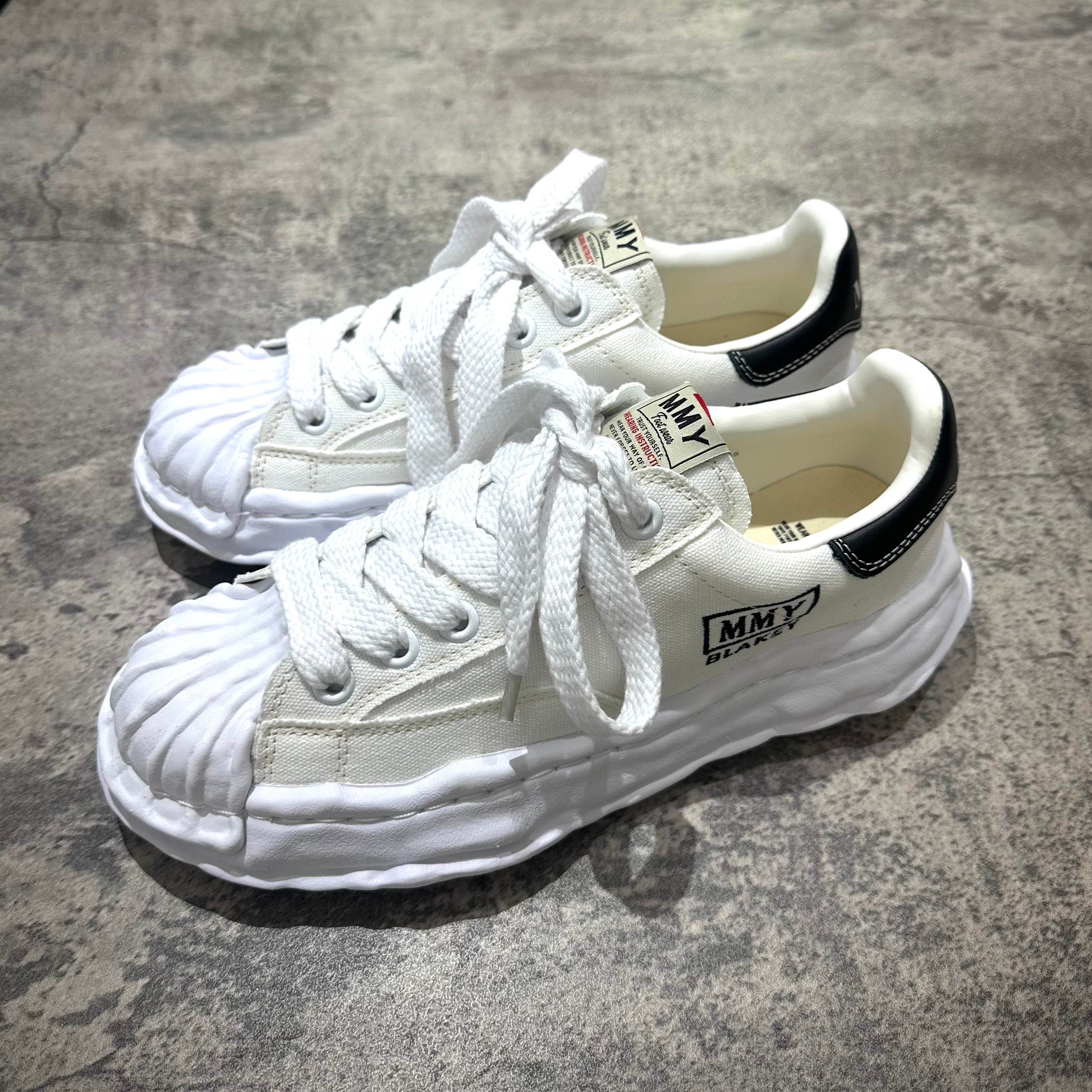 MMY BLAKEY OG Canvas - White sneakers with unique design and black accents on a textured floor.