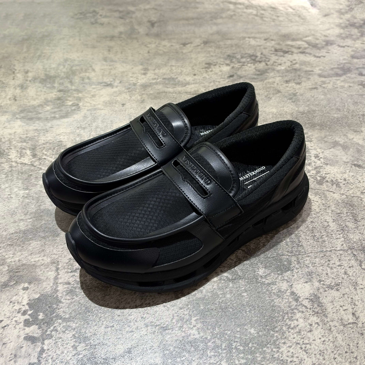 Mastermind World x Moisthrough 360 Hobalips black slip-on shoes on a concrete floor.