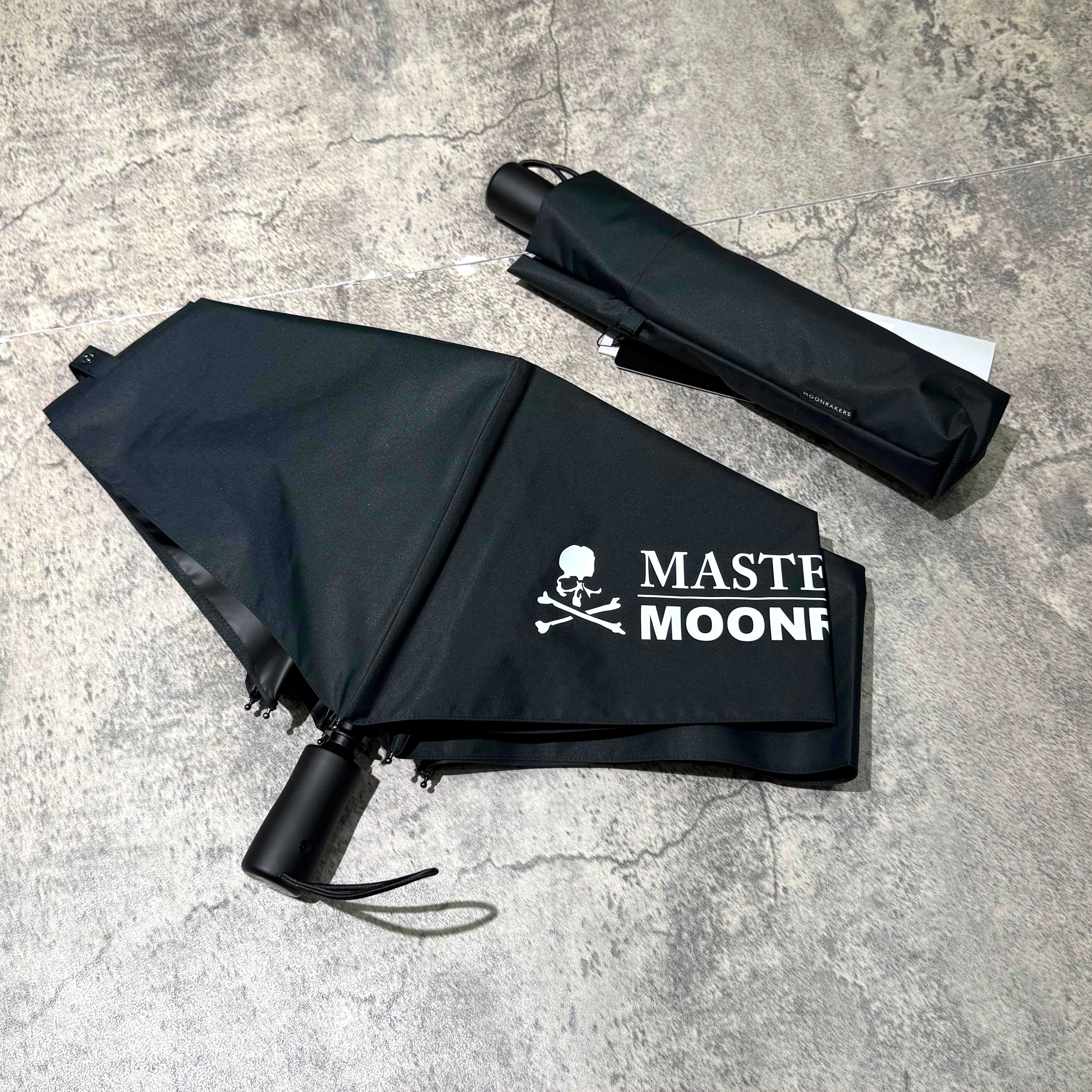 Mastermind Japan Moonrakers Umbrella in black with logo and compact case on a textured surface.